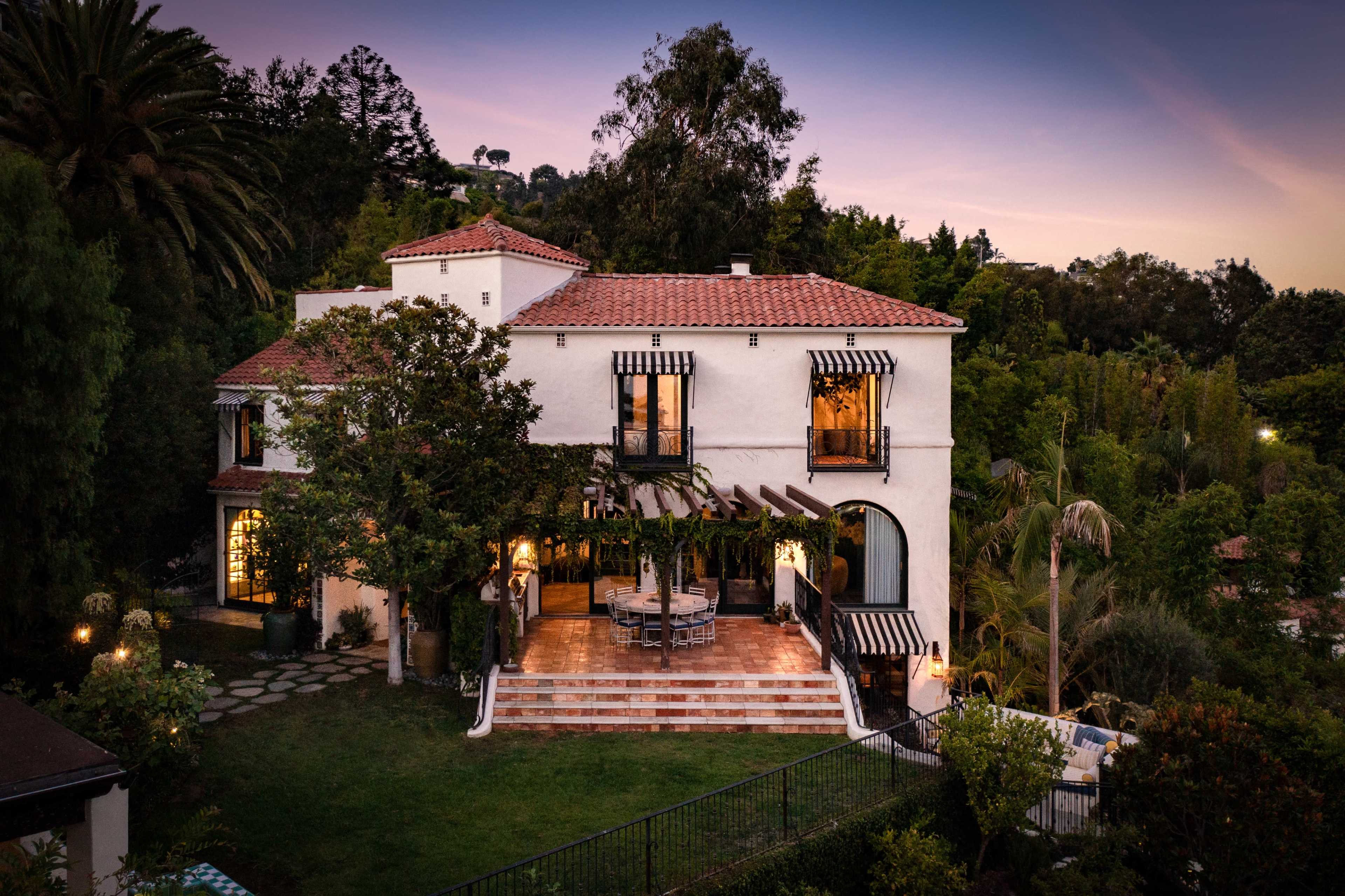 A two-story white house with a terracotta roof is surrounded by lush greenery and features a large outdoor patio with a dining area.