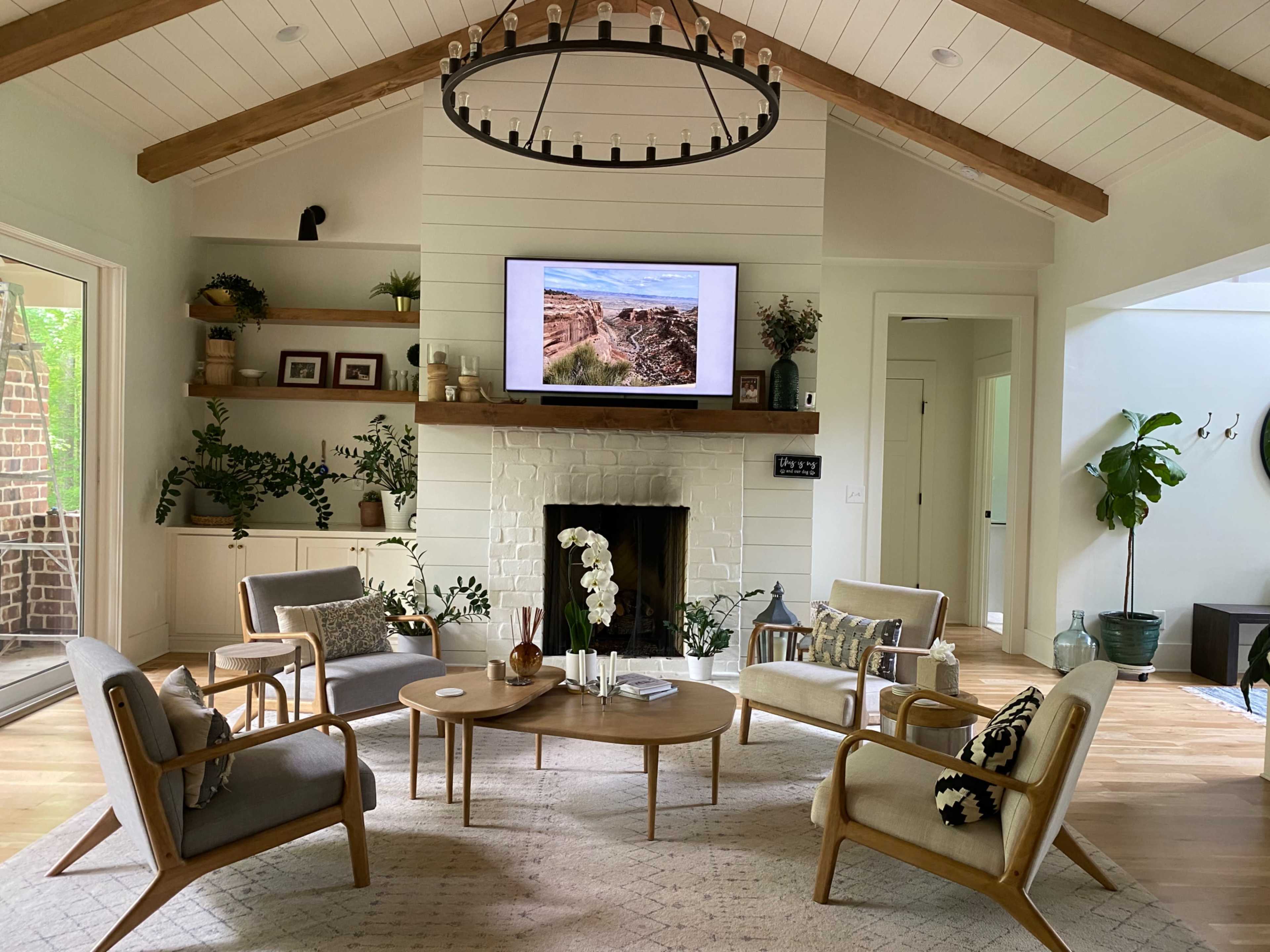 A cozy living room features a central coffee table surrounded by four chairs, a large television mounted above a white brick fireplace, and decor elements like plants and framed pictures on the walls.