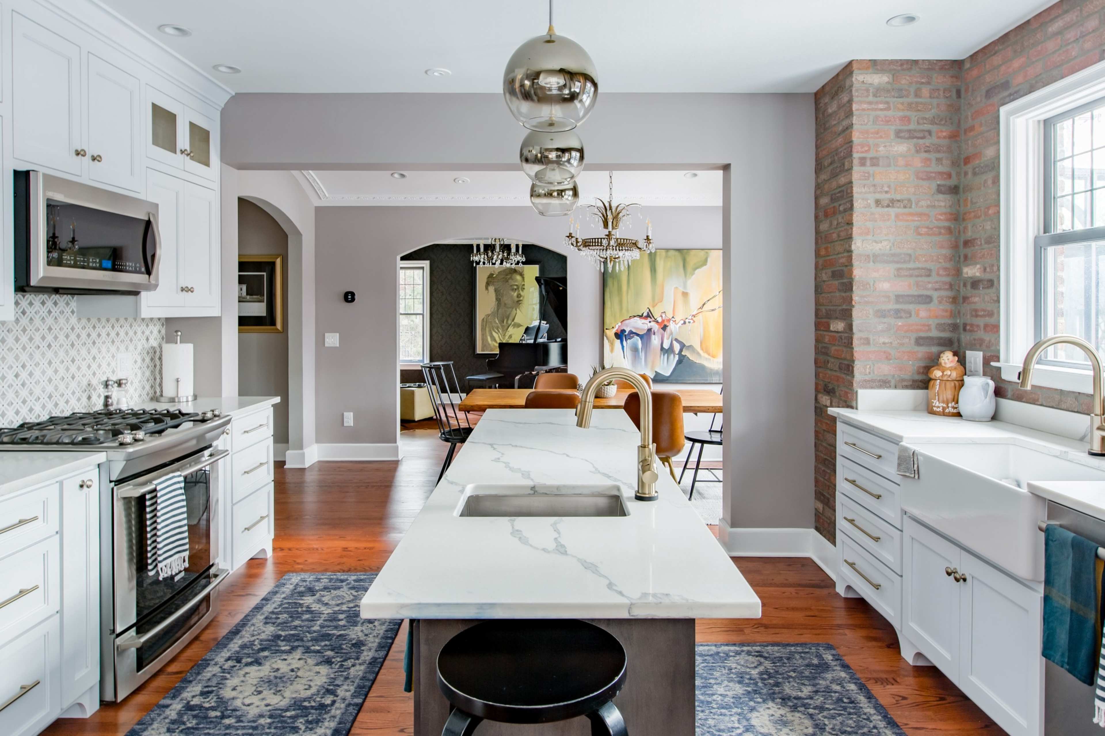 A modern kitchen with white cabinetry, a central island with a marble countertop, and exposed brick walls adjacent to a dining area.