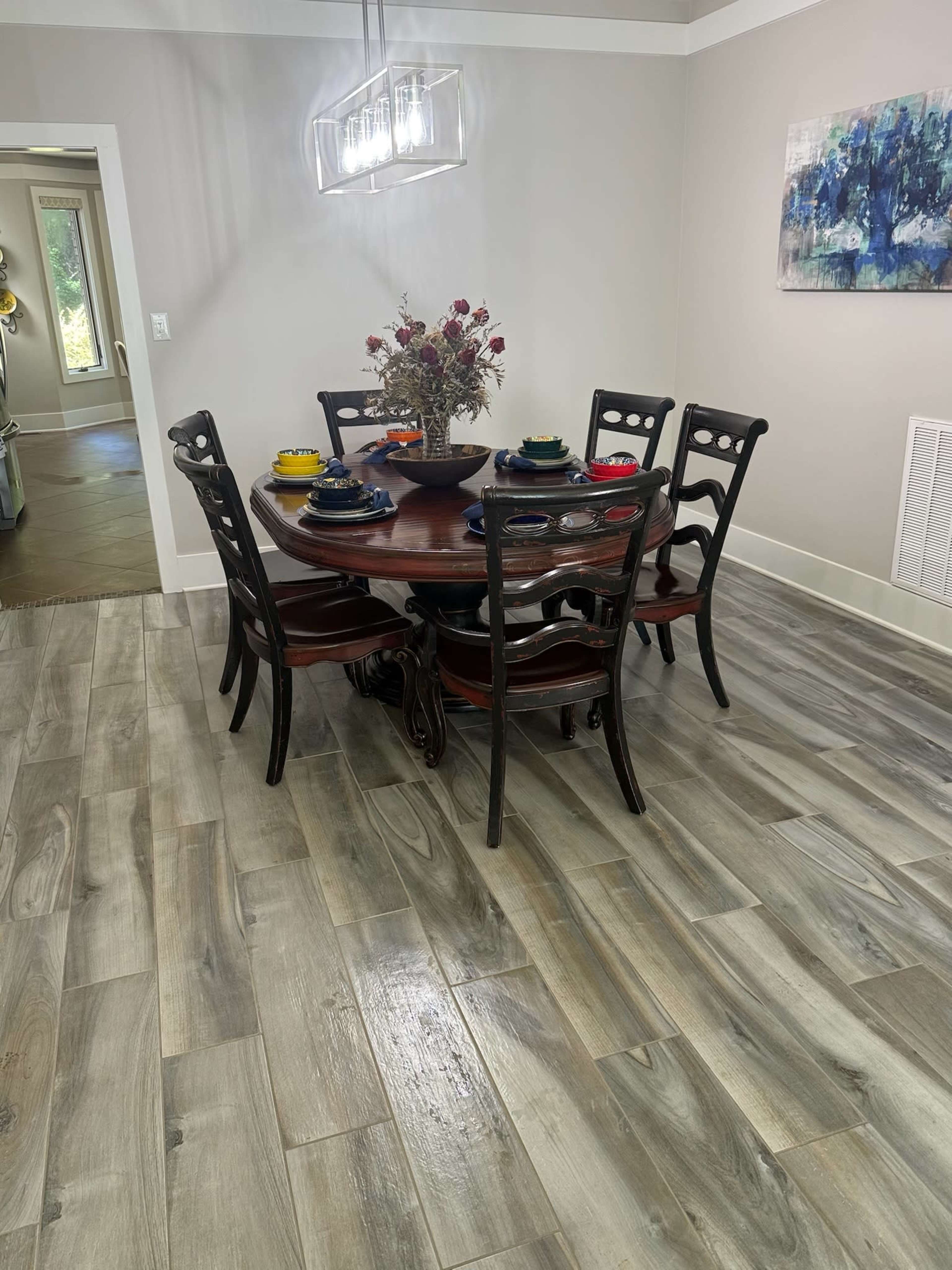 A round wooden dining table is set with plates and utensils, surrounded by six black chairs in a room with gray tile flooring and a decorative centerpiece.
