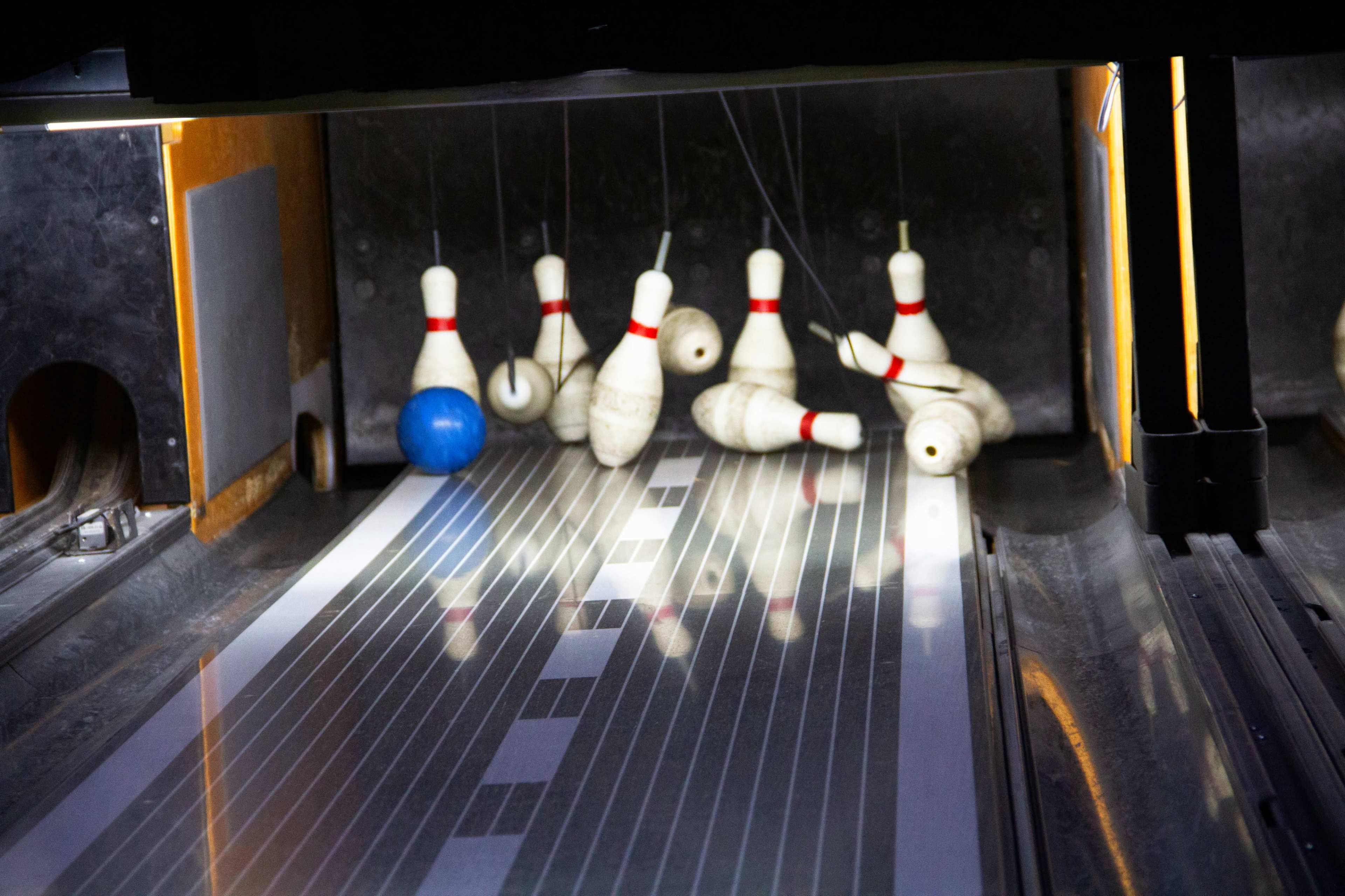 A blue bowling ball rolls down a wooden lane toward a set of white pins, some of which are swaying slightly.