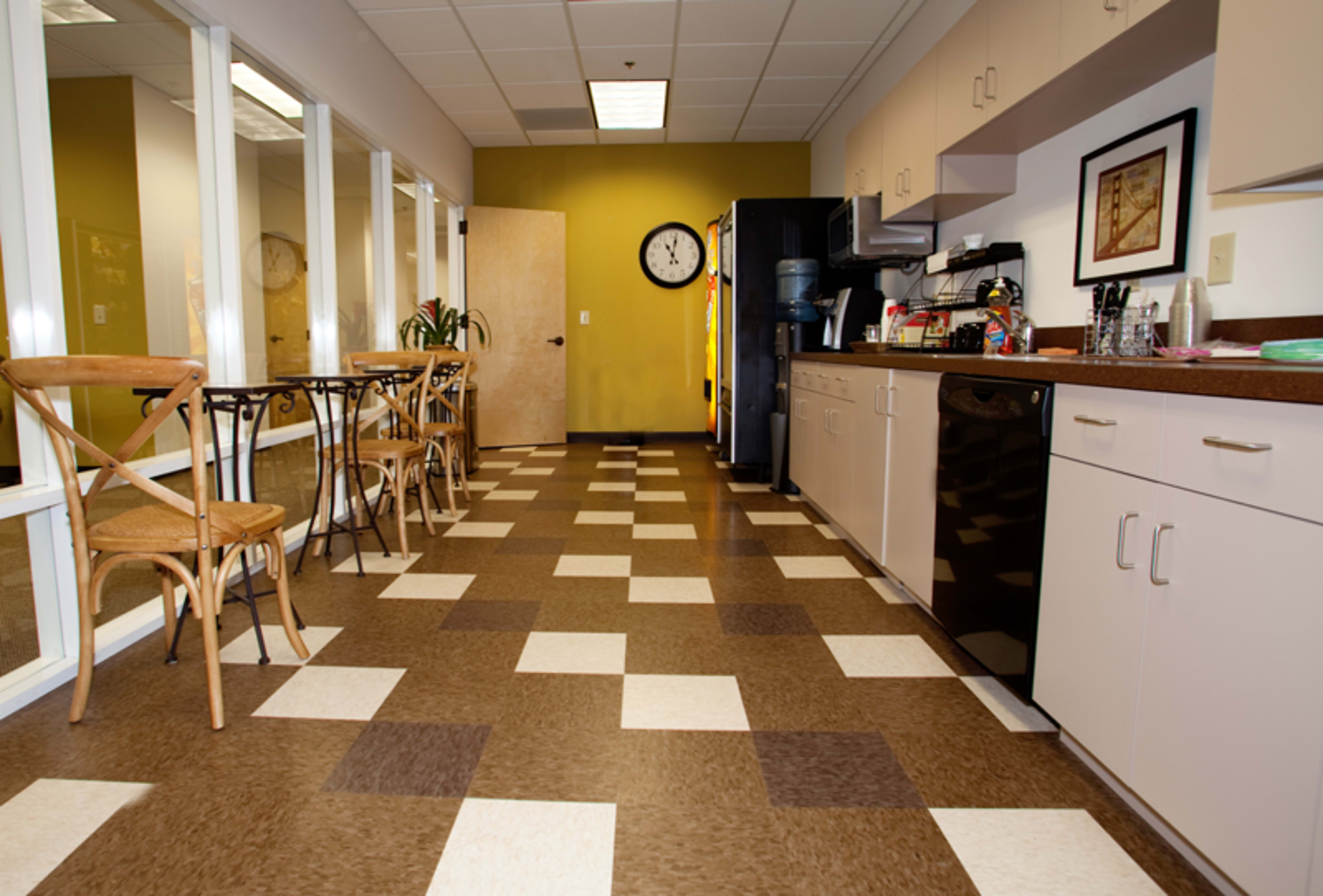 A break room with a tiled floor, a kitchenette along one wall, a dining table with chairs, and large windows on the opposite side.