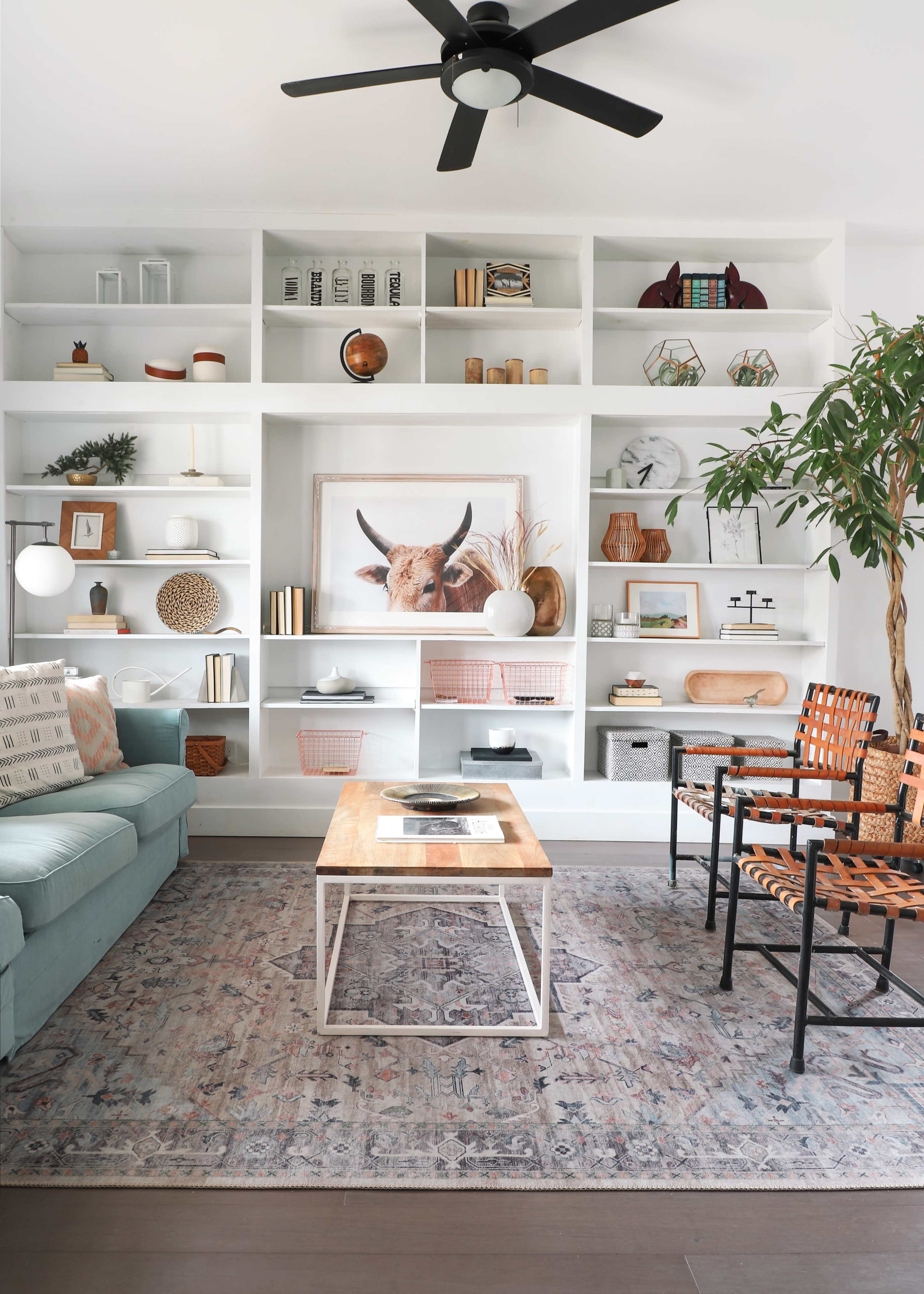 A modern living room features a light blue sofa, a wooden coffee table, and a wall of white shelves displaying decorative items and art.