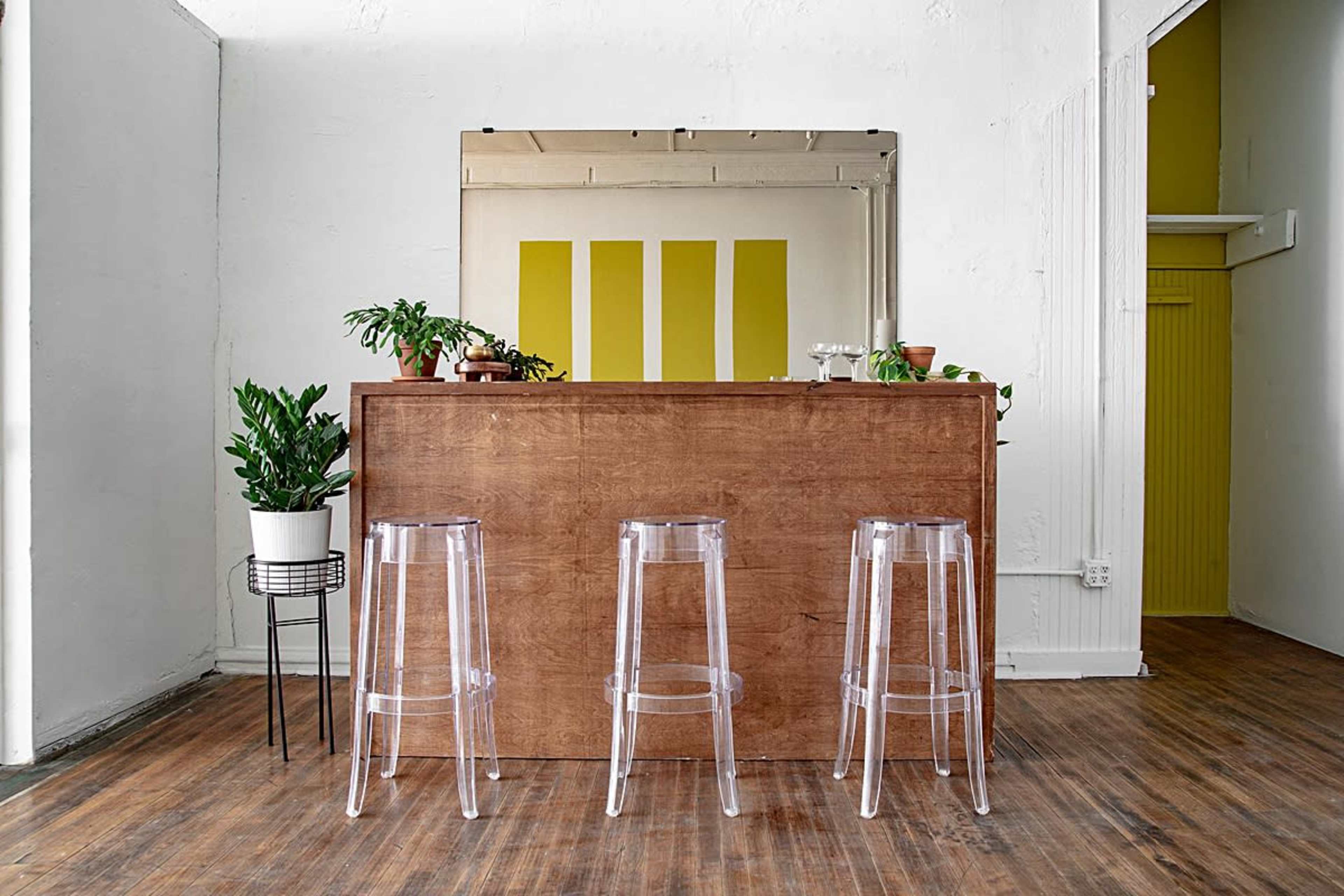 A wooden bar with three clear stools and potted plants is set against a wall with a yellow-striped mural.