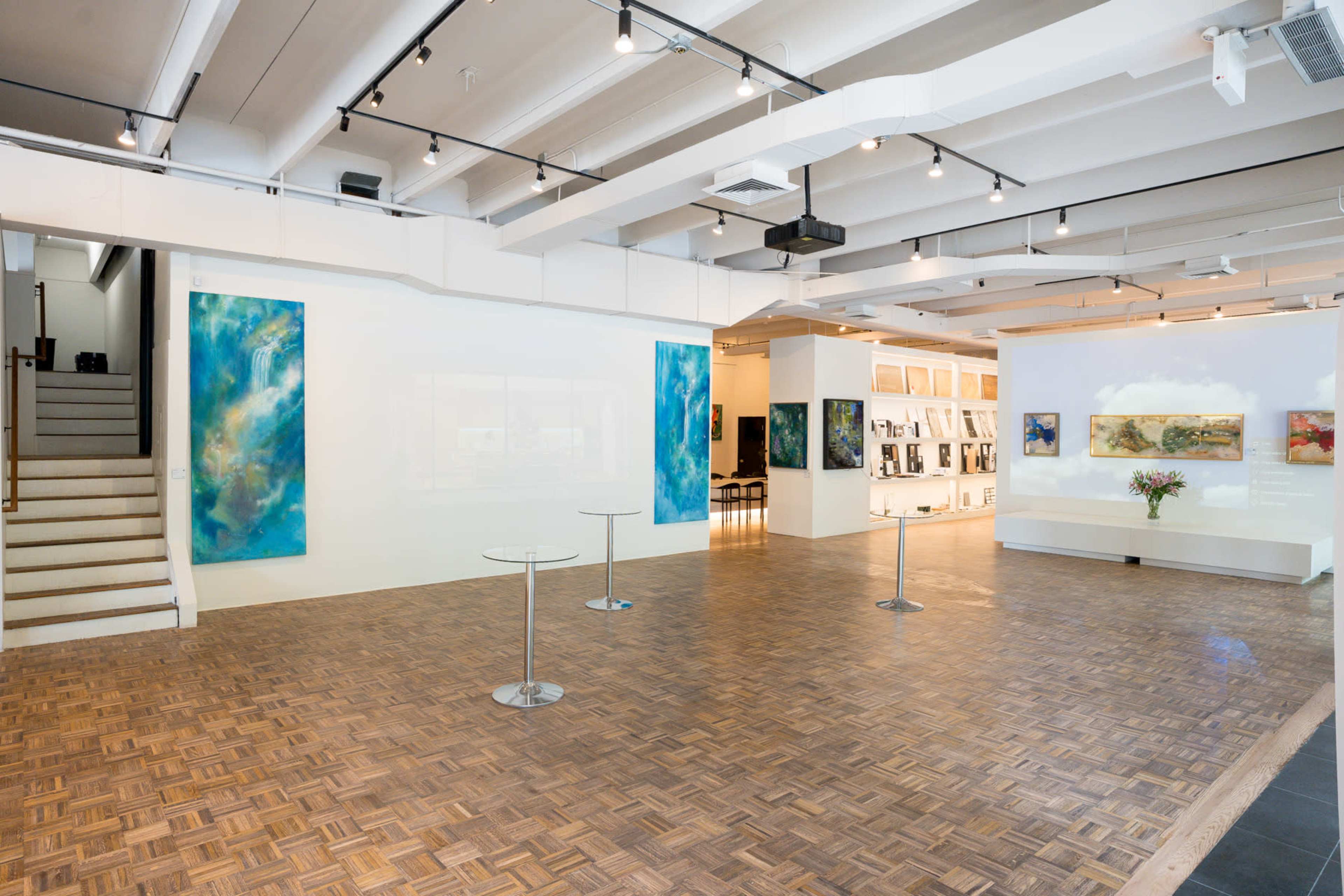 The image depicts a spacious art gallery featuring wooden flooring, vibrant abstract paintings on the walls, and minimalist glass display tables.