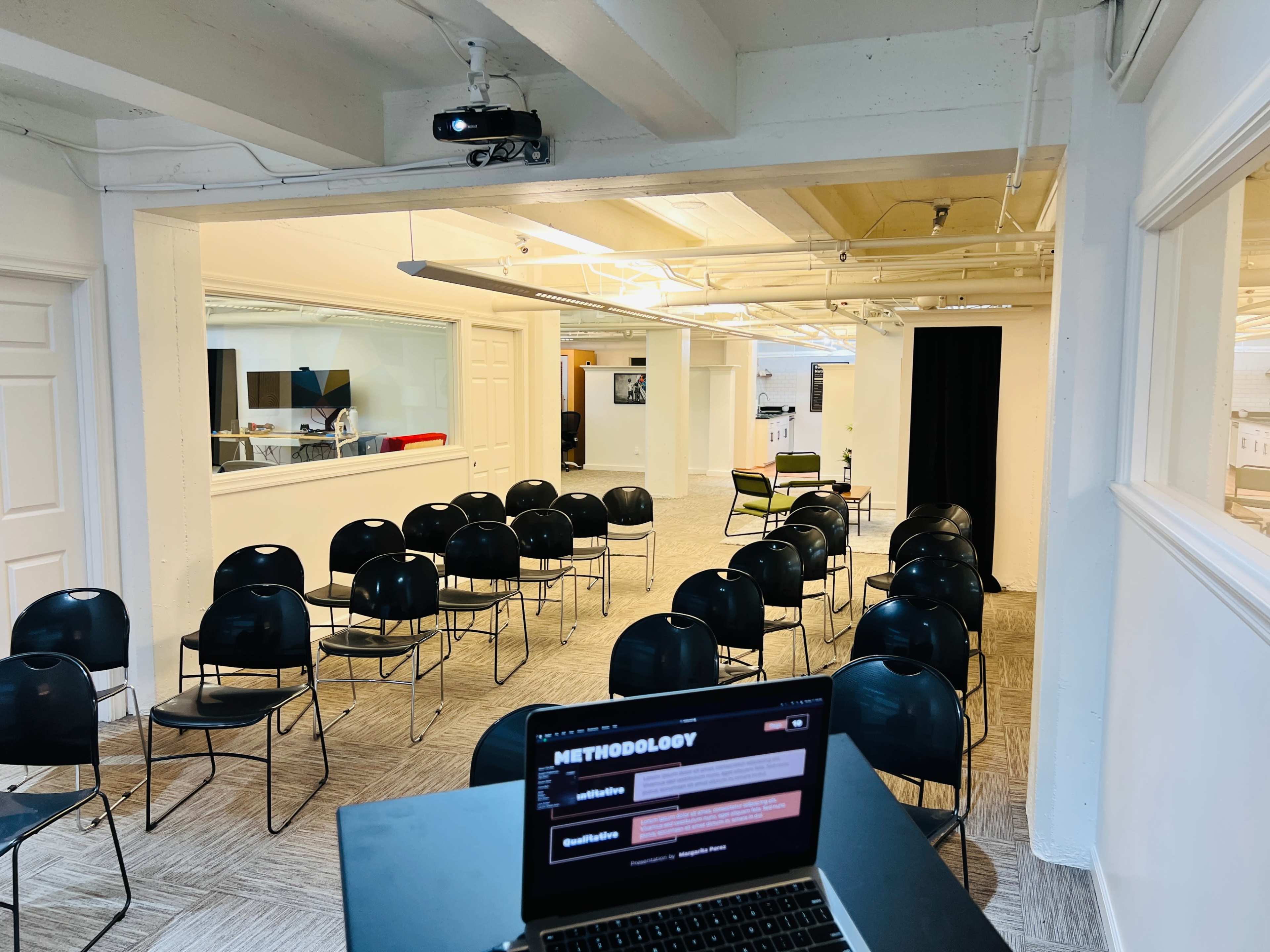 A spacious room with rows of black chairs facing a projector screen and a laptop on a table in the foreground.