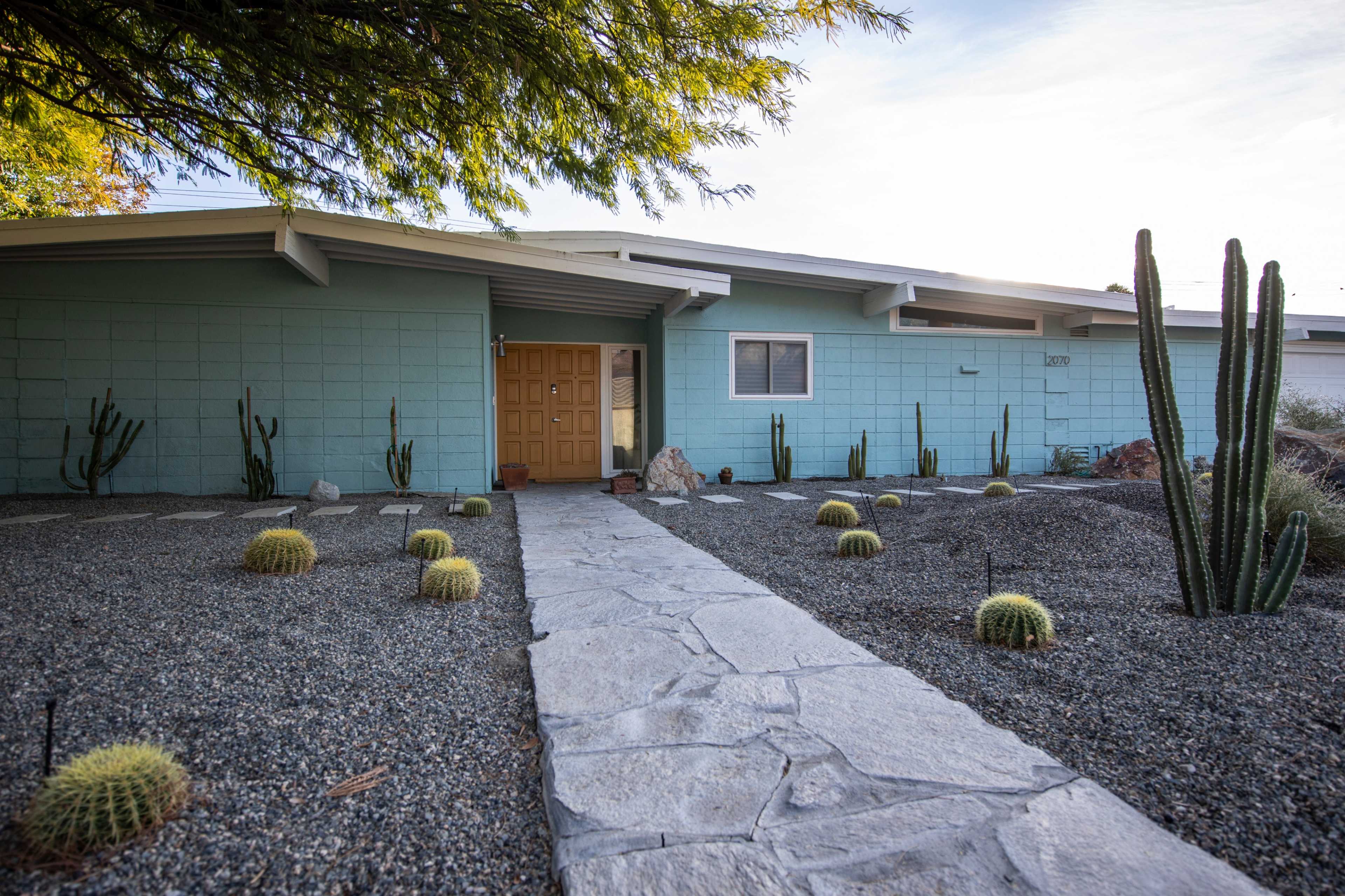 A modern home with a light blue exterior and a stone pathway leading to a wooden door, surrounded by a landscaped area featuring cacti and gravel.