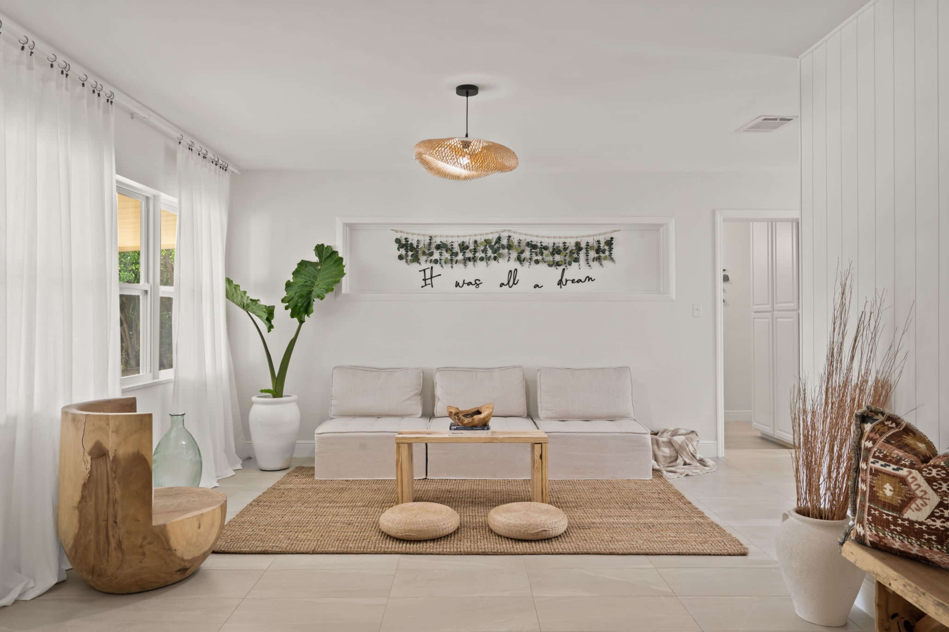 A living room featuring a light-colored couch, a wooden coffee table, two circular ottomans, and decor elements like a large plant and wall artwork.