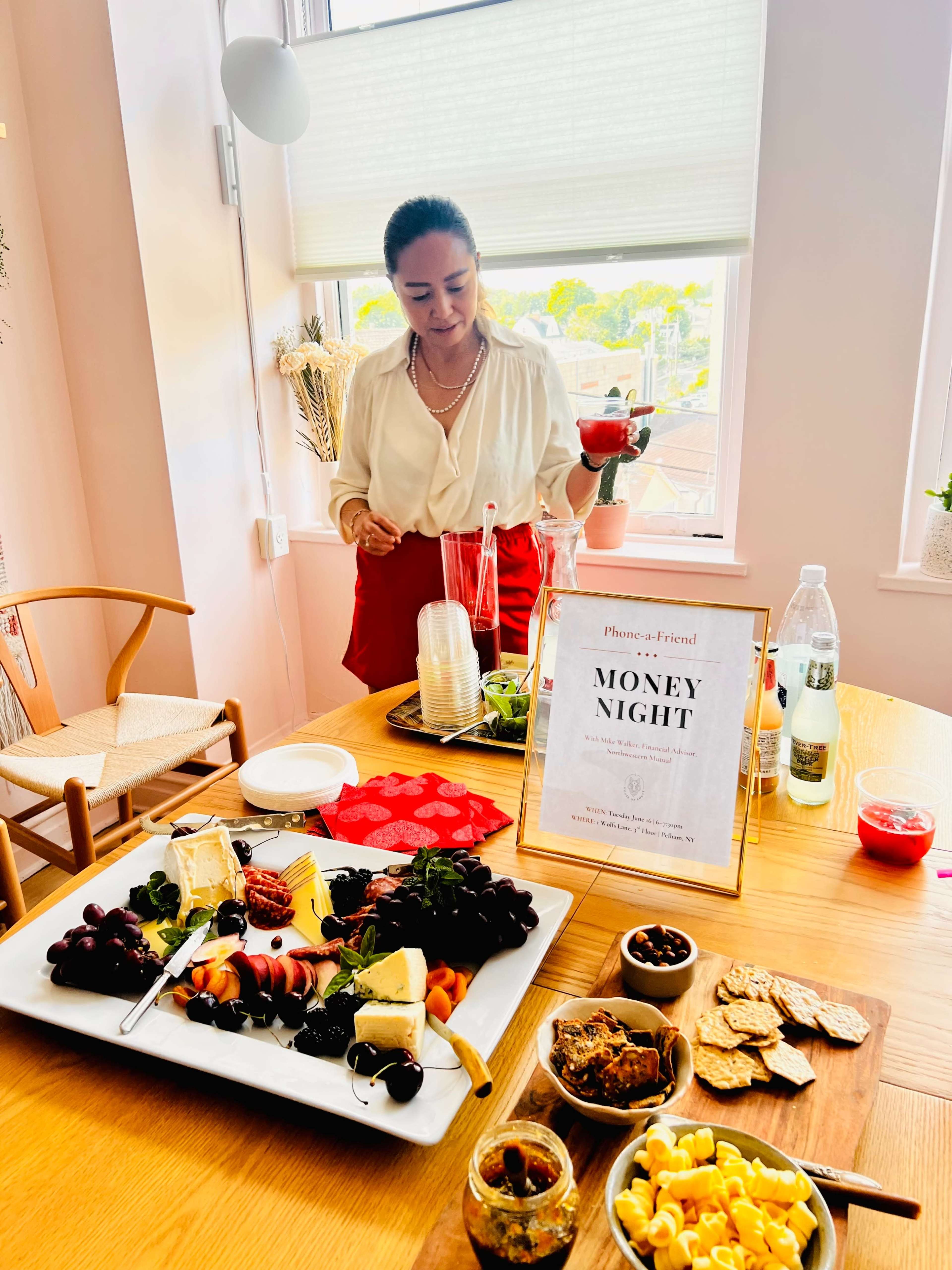 A woman in a white blouse and red skirt stands beside a table featuring a variety of food items, including a cheese platter and snacks, while a sign that reads "MONEY NIGHT" is displayed on the table.