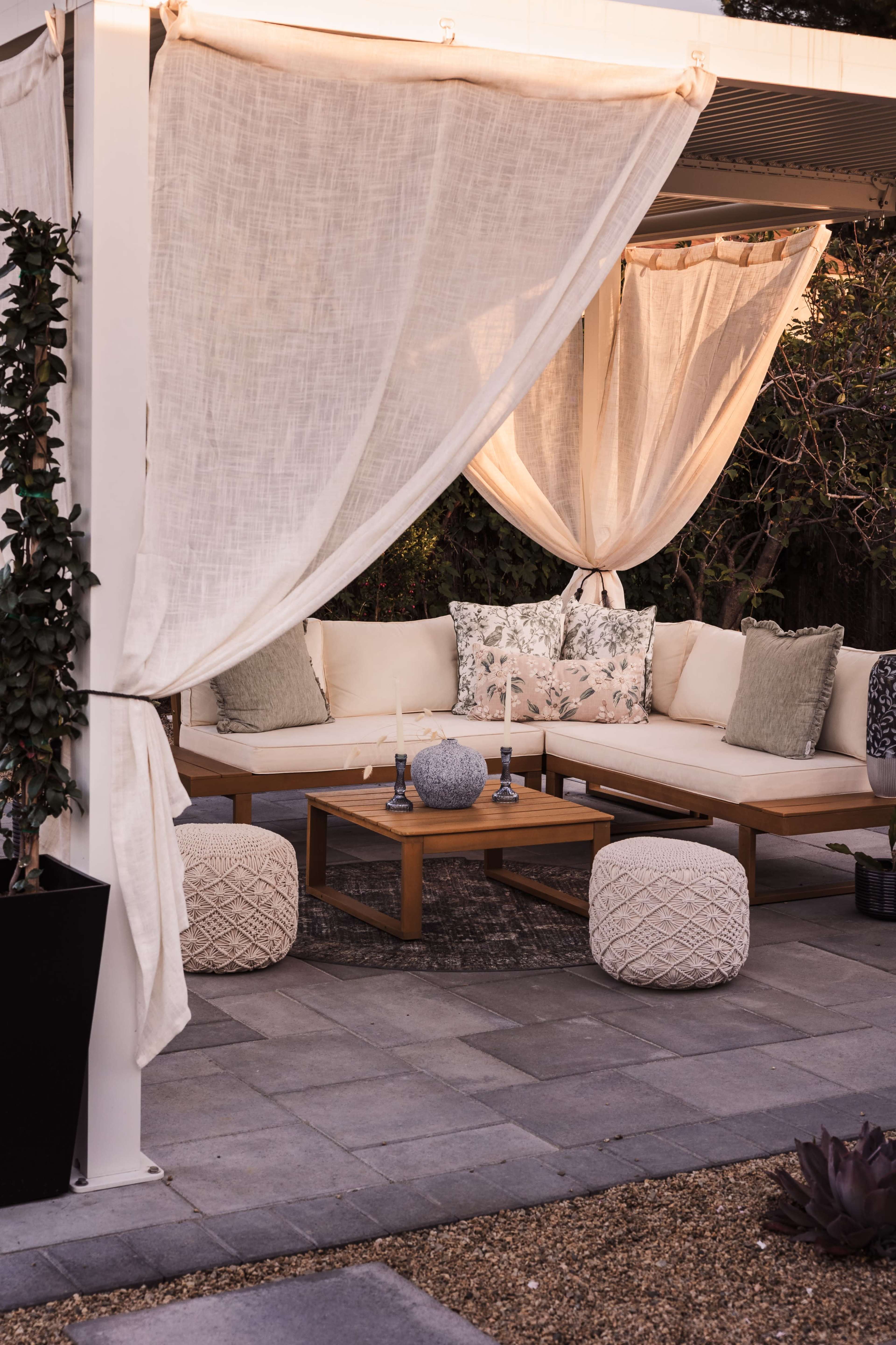 A cozy outdoor seating area features a cream-colored sectional sofa with decorative cushions, a wooden coffee table, and soft lighting from candles.