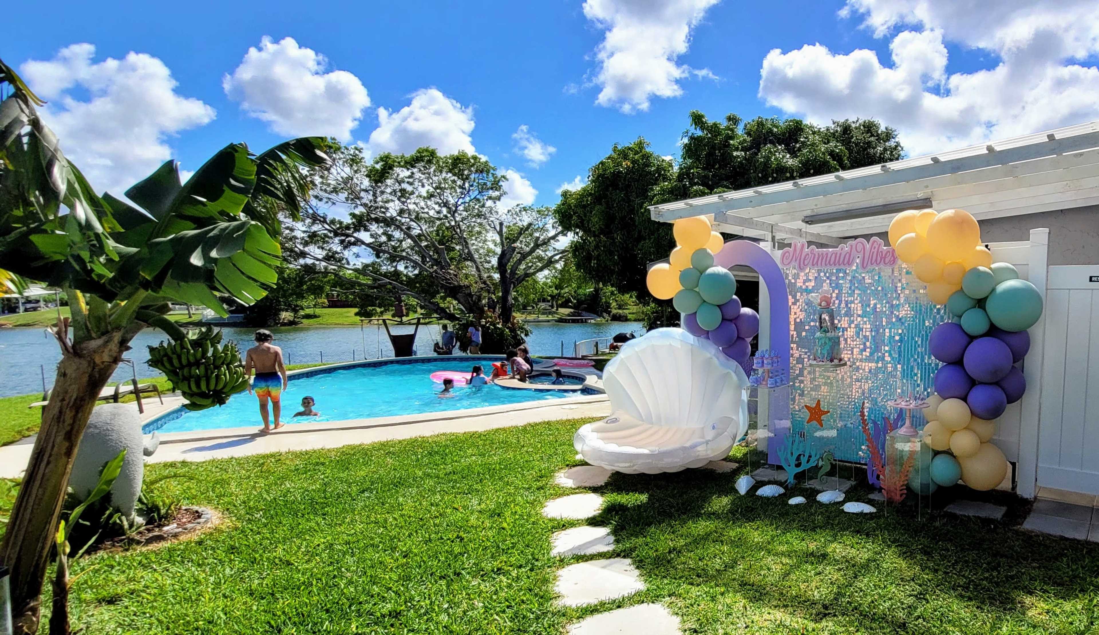 A backyard scene features a swimming pool with children playing, surrounded by lush greenery and decorated with a themed display of balloons and a large shell.
