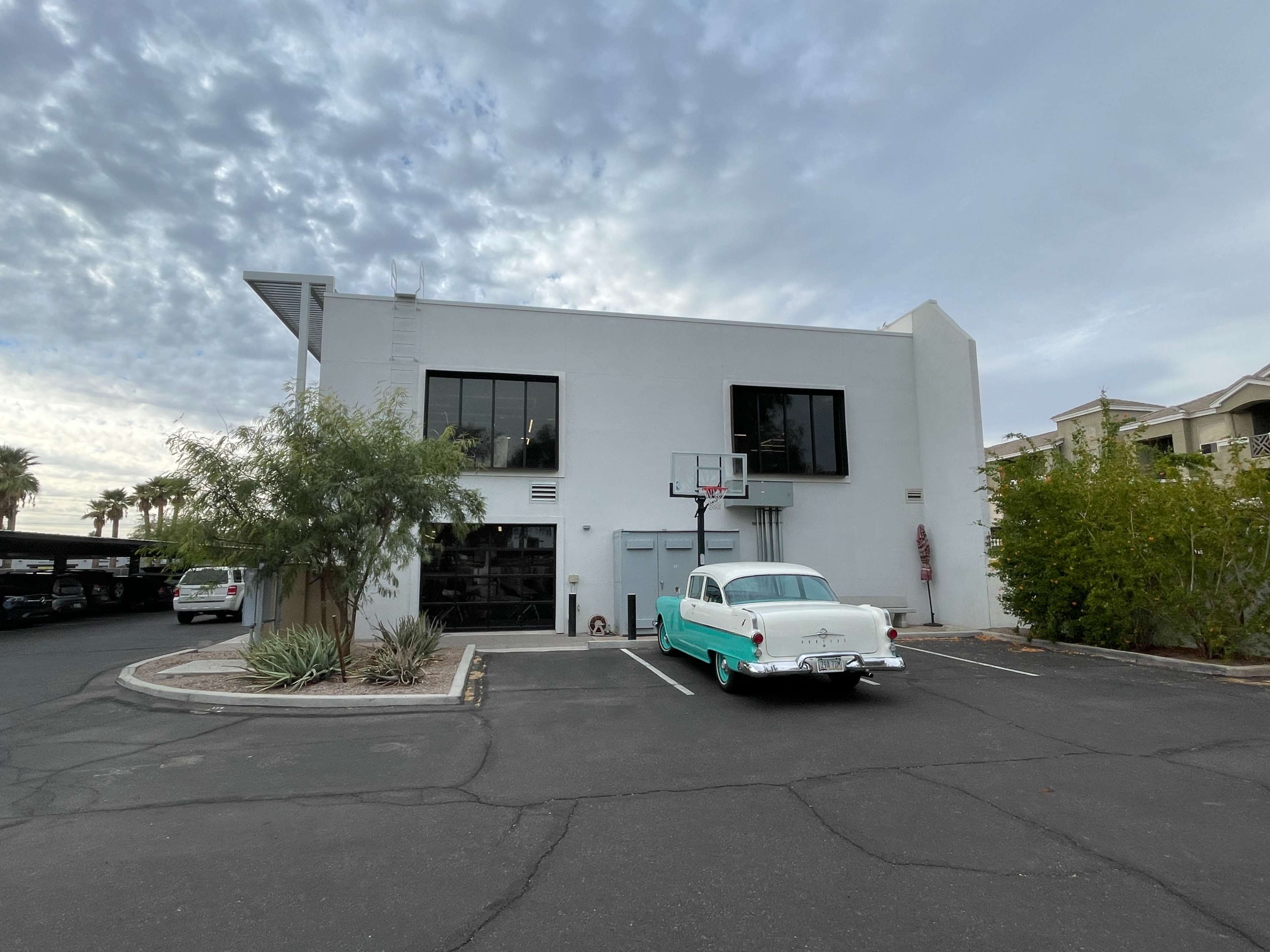 The image shows a vintage turquoise car parked in front of a modern white building with large windows and a basketball hoop.