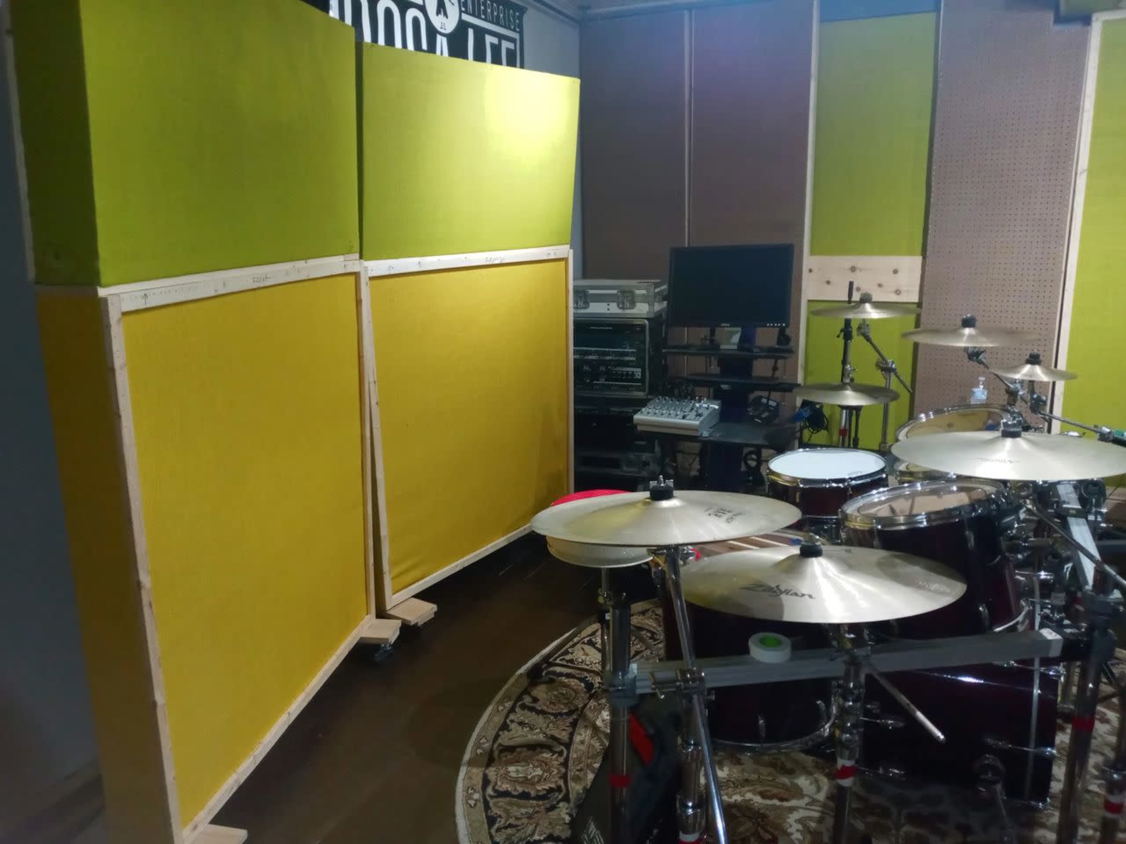 The image shows a section of a music studio featuring a drum set, large green acoustic panels, and various audio equipment in the background.