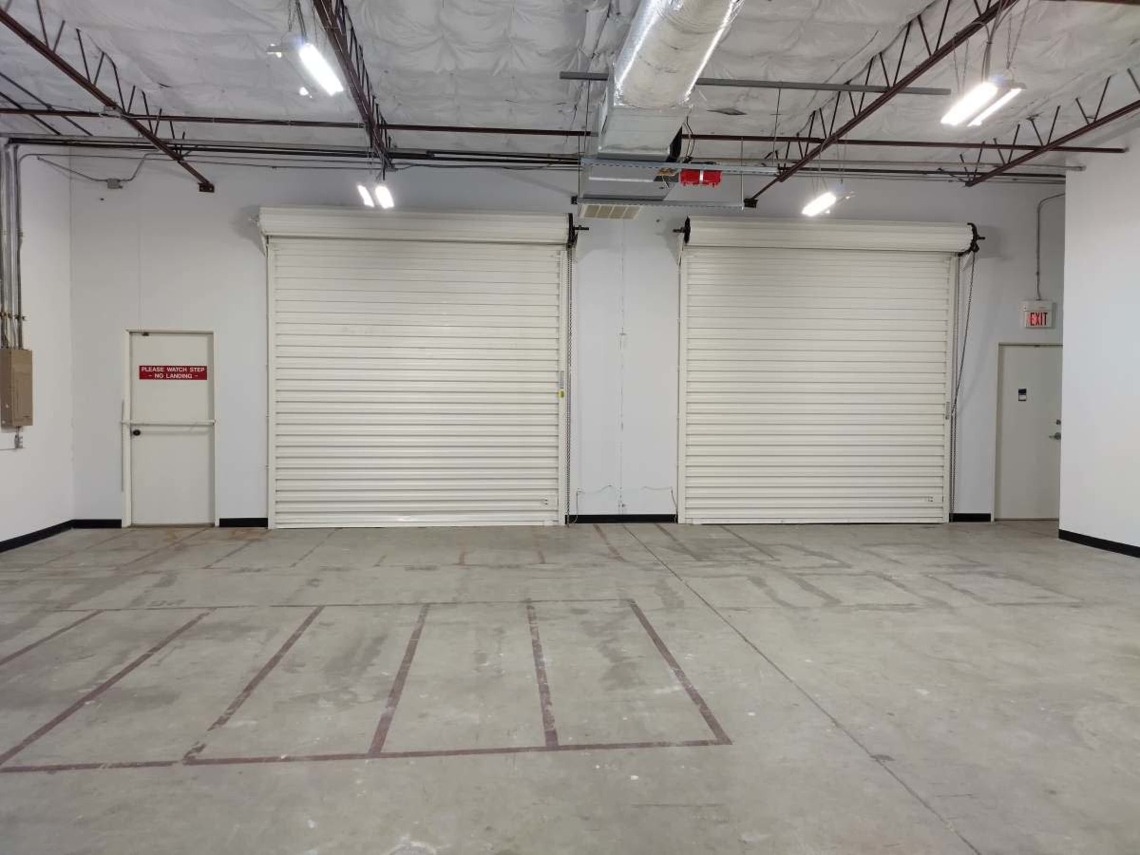 The image shows an empty industrial space with two large white roll-up doors and a door on the left side.