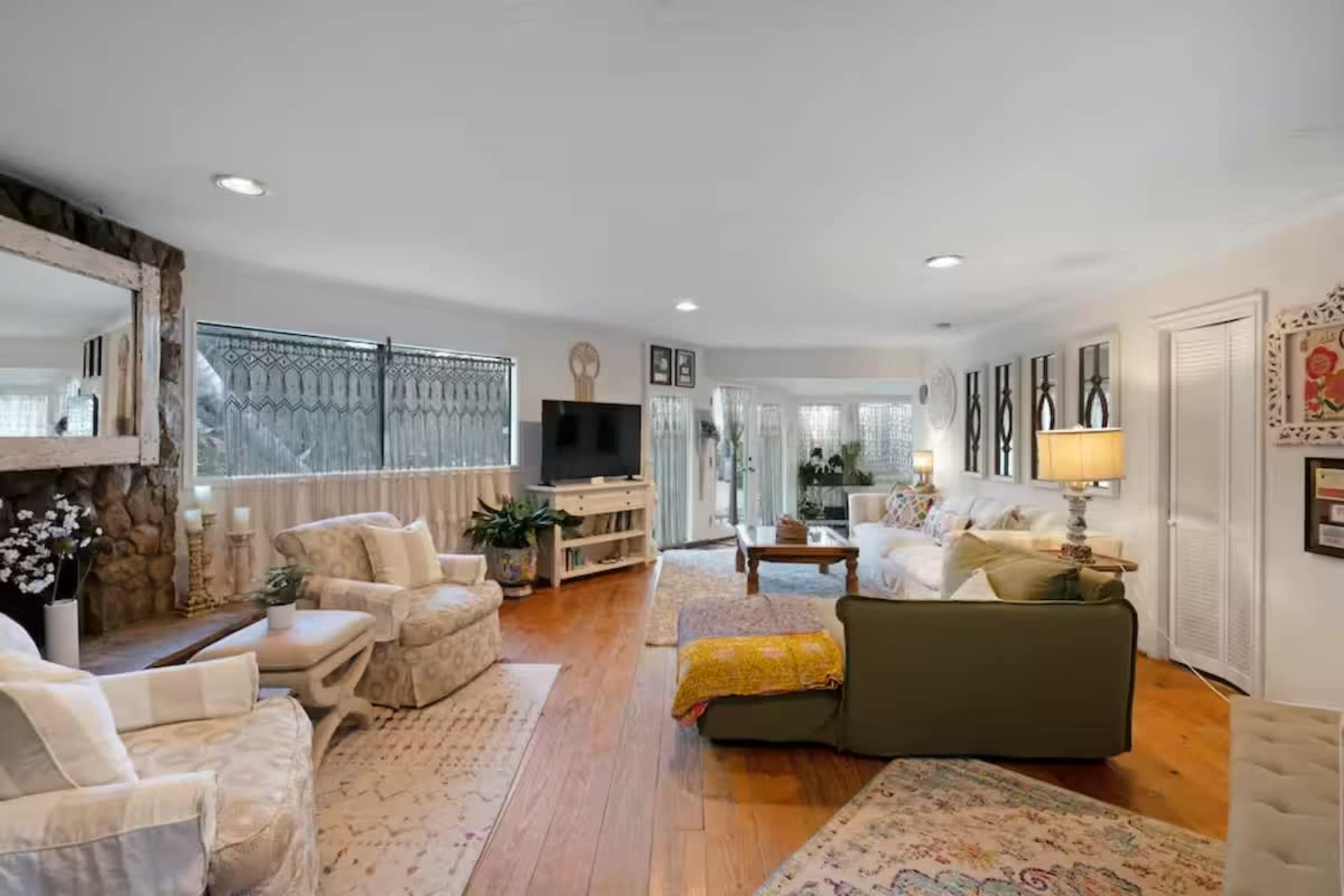 The image shows a spacious living room with hardwood floors, a stone fireplace, and large windows letting in natural light, furnished with various seating options and decorative elements.