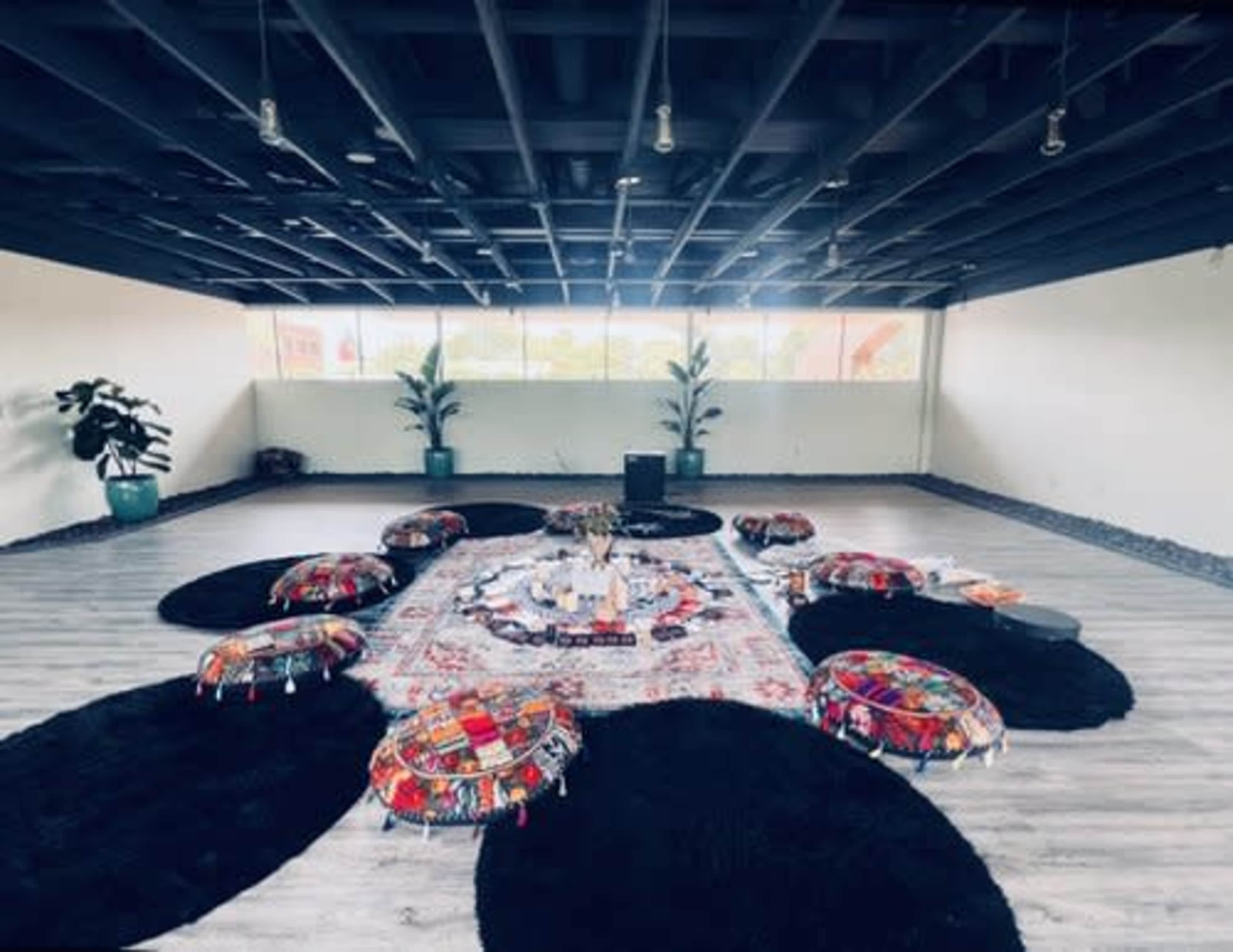 The image shows a spacious, minimalist room with a centrally arranged area featuring colorful poufs and a decorative rug surrounded by plants.