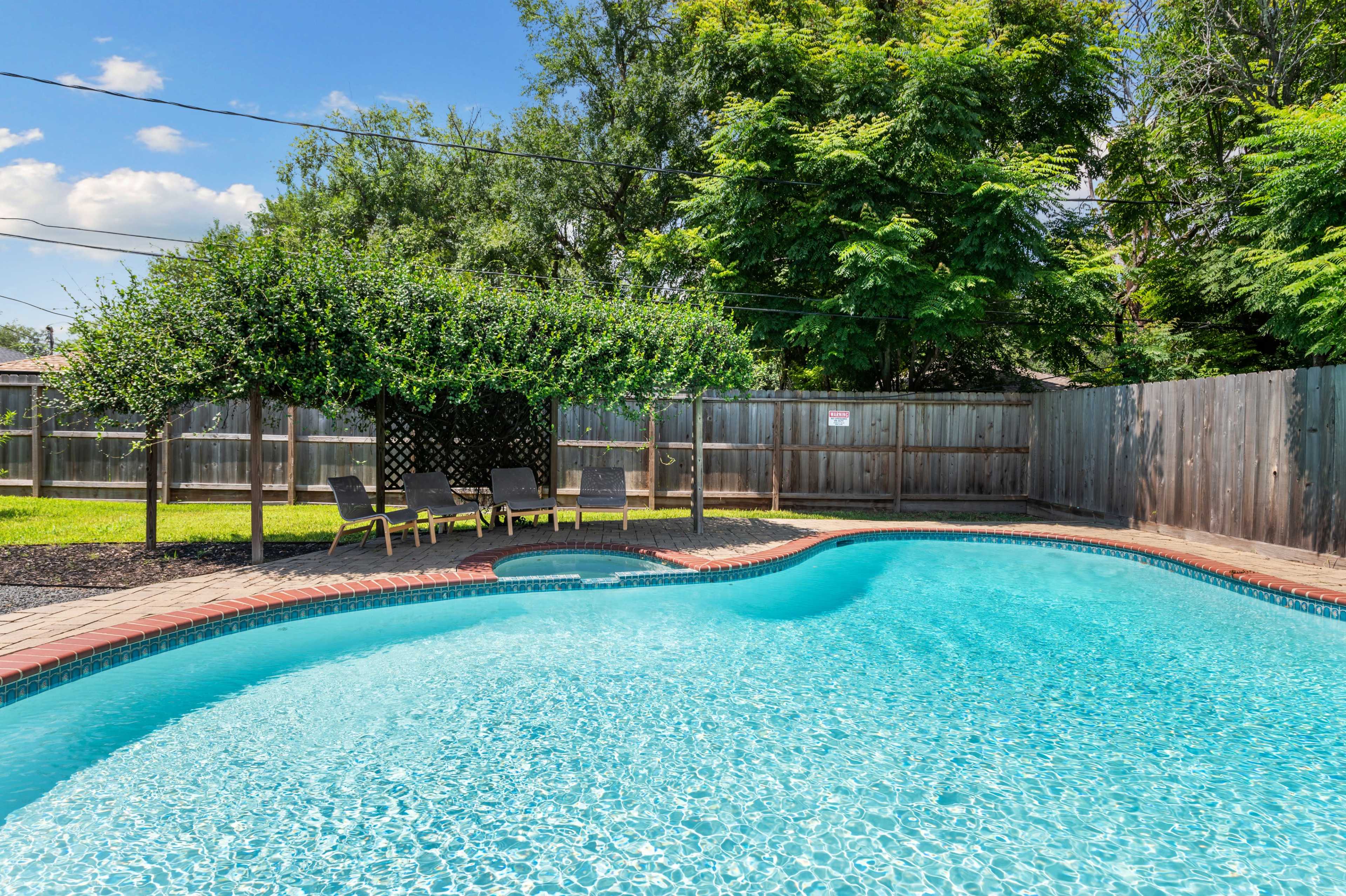 A residential backyard with a clear blue swimming pool surrounded by lounge chairs and a shaded area created by trees.