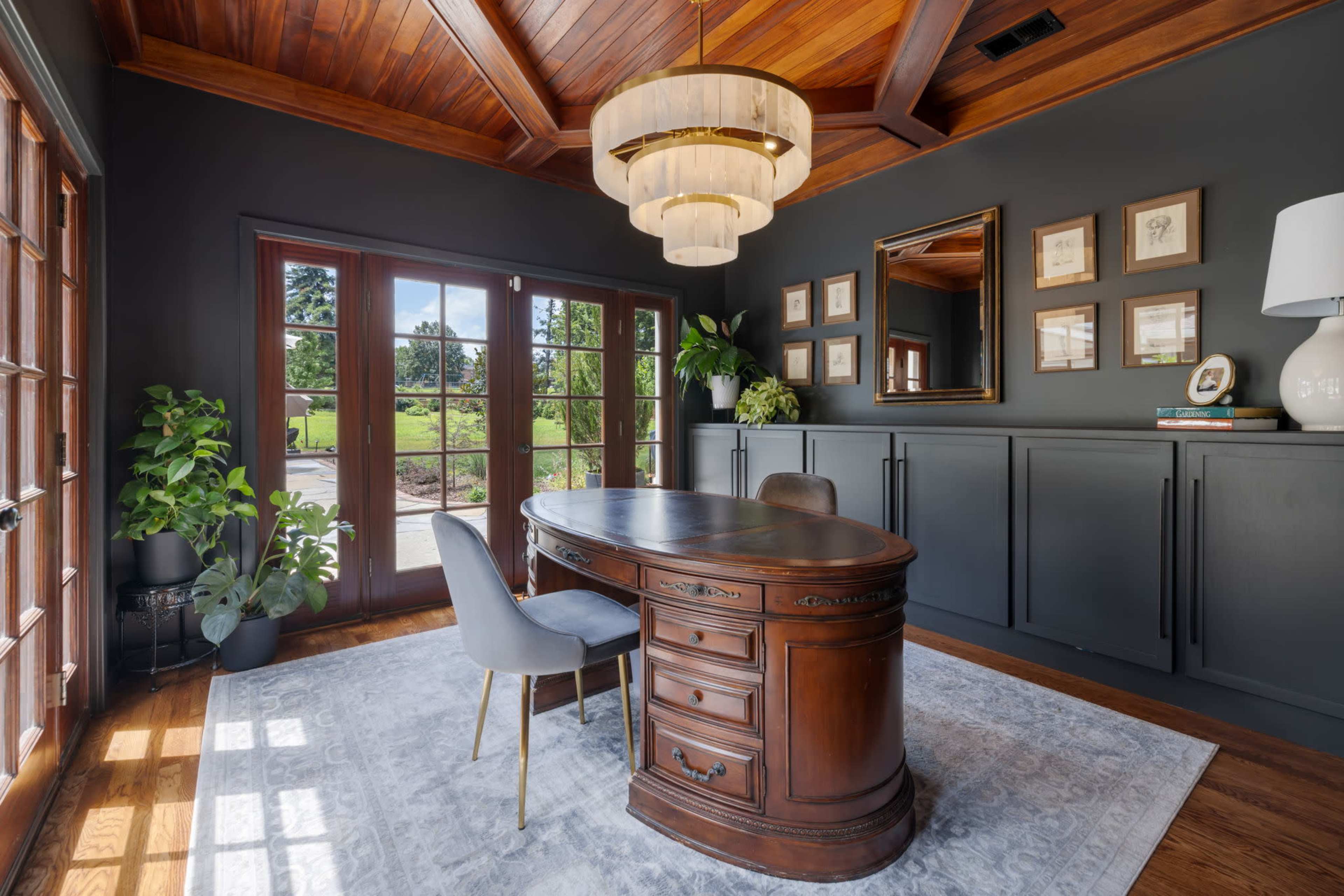The image shows a home office with a wooden desk, a gray chair, large windows revealing a view of greenery, and modern decor elements.