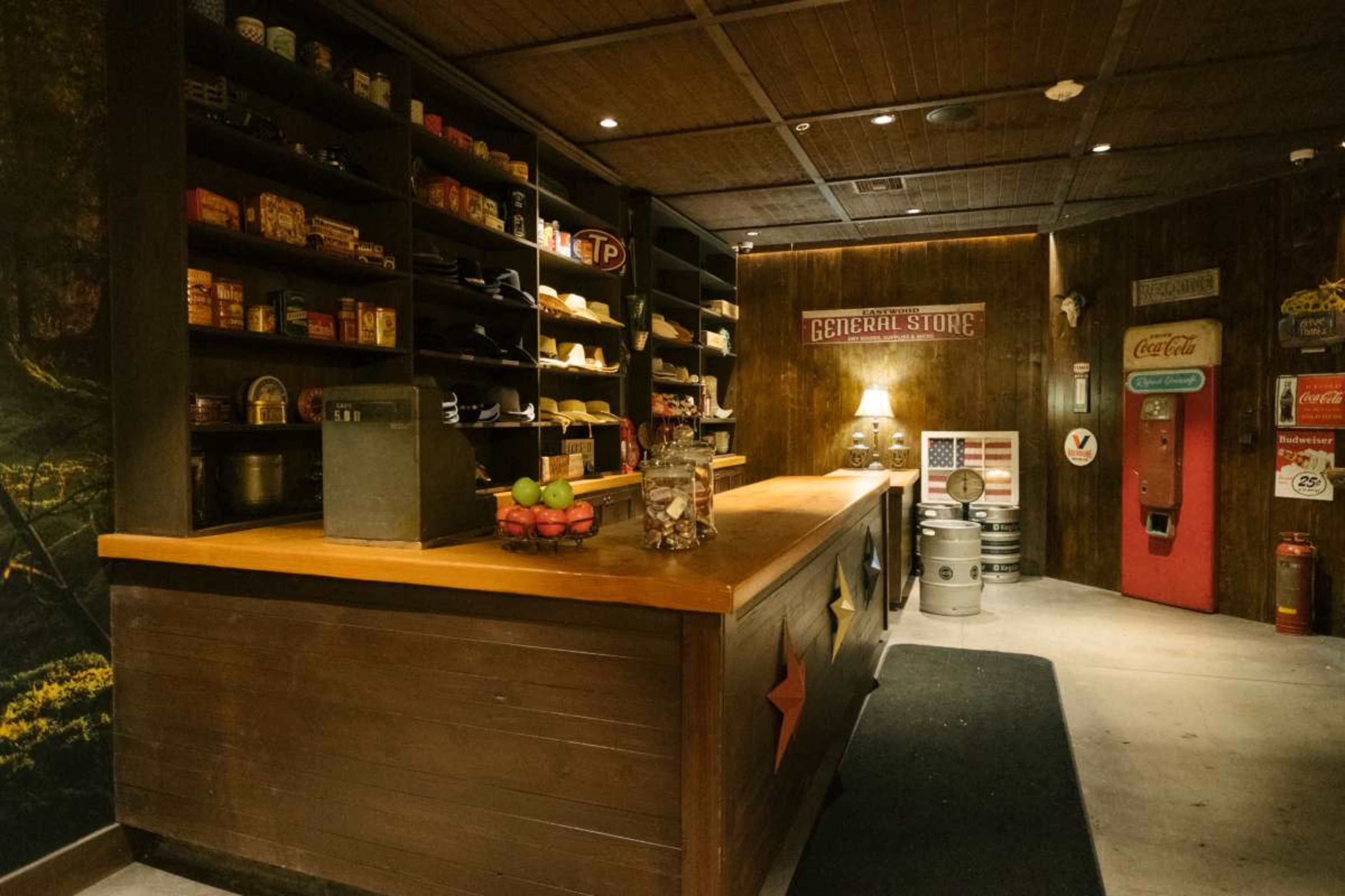 The image shows a rustic general store interior with wooden shelves stocked with canned goods and a wooden counter displaying fruit and a cash register.