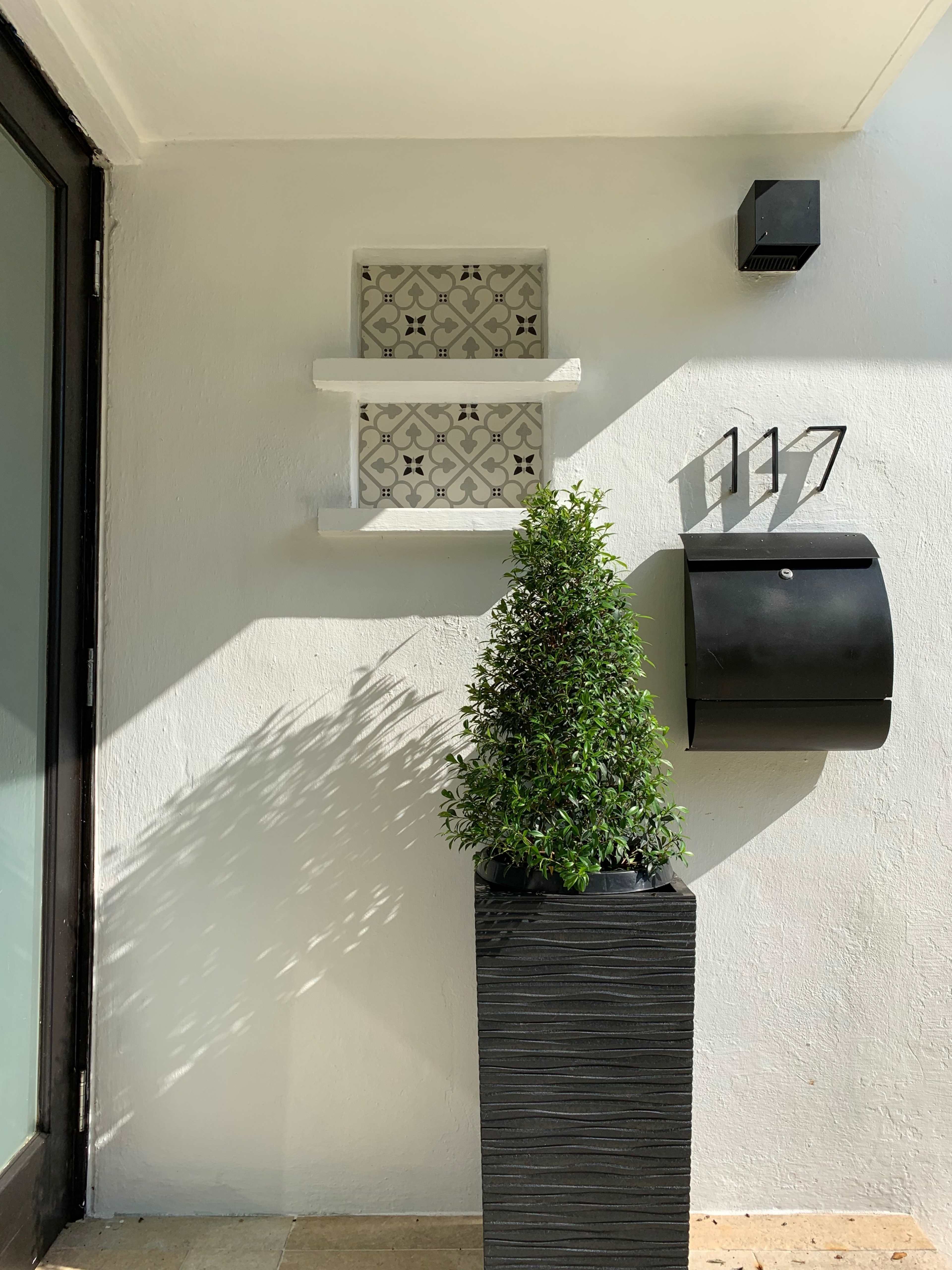 The image shows a planter with a neatly trimmed shrub beside a decorative window and a black mailbox mounted on a white wall.