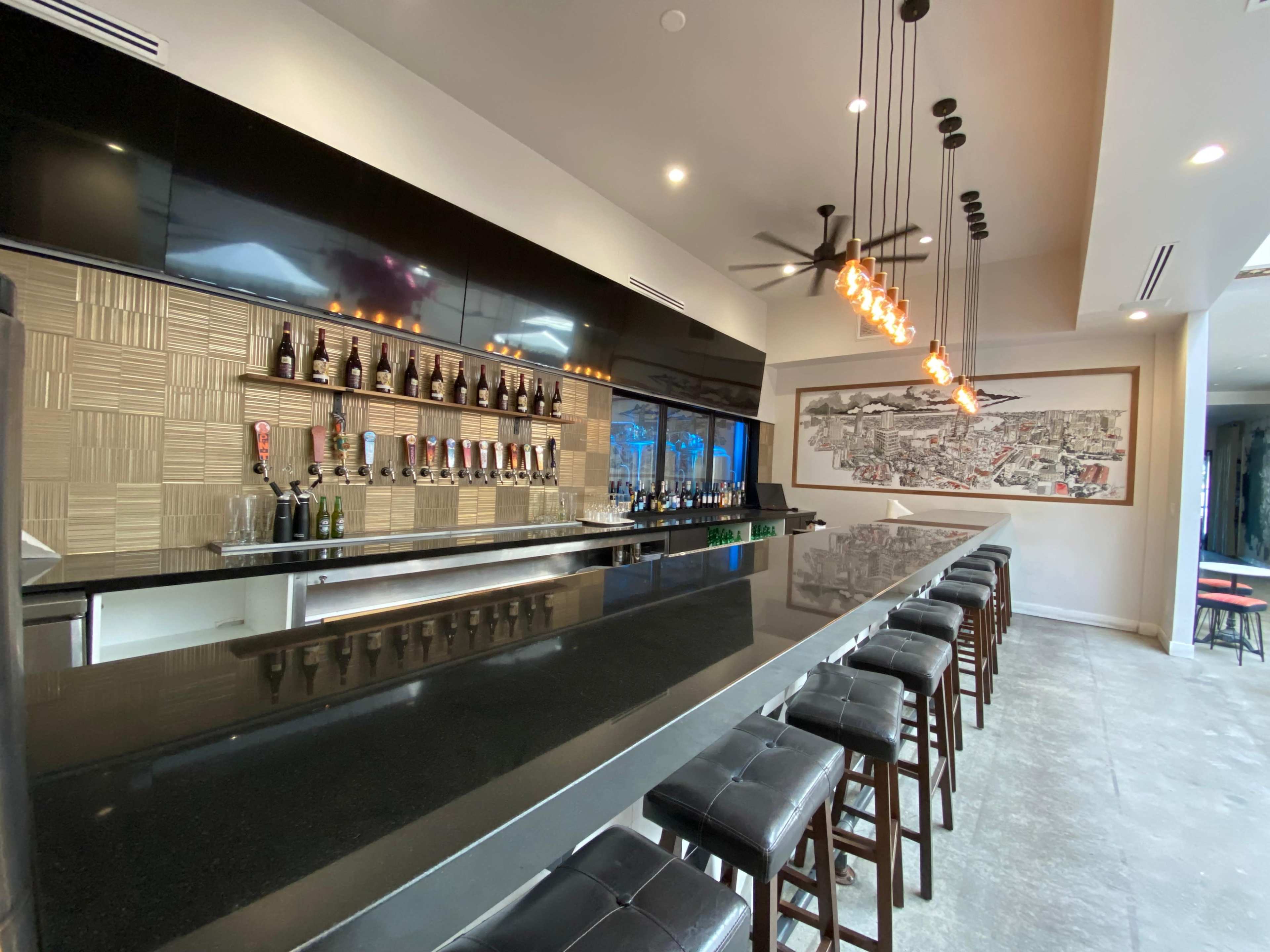 A modern bar features a long black countertop, wooden stools, and shelves lined with various bottles behind it.