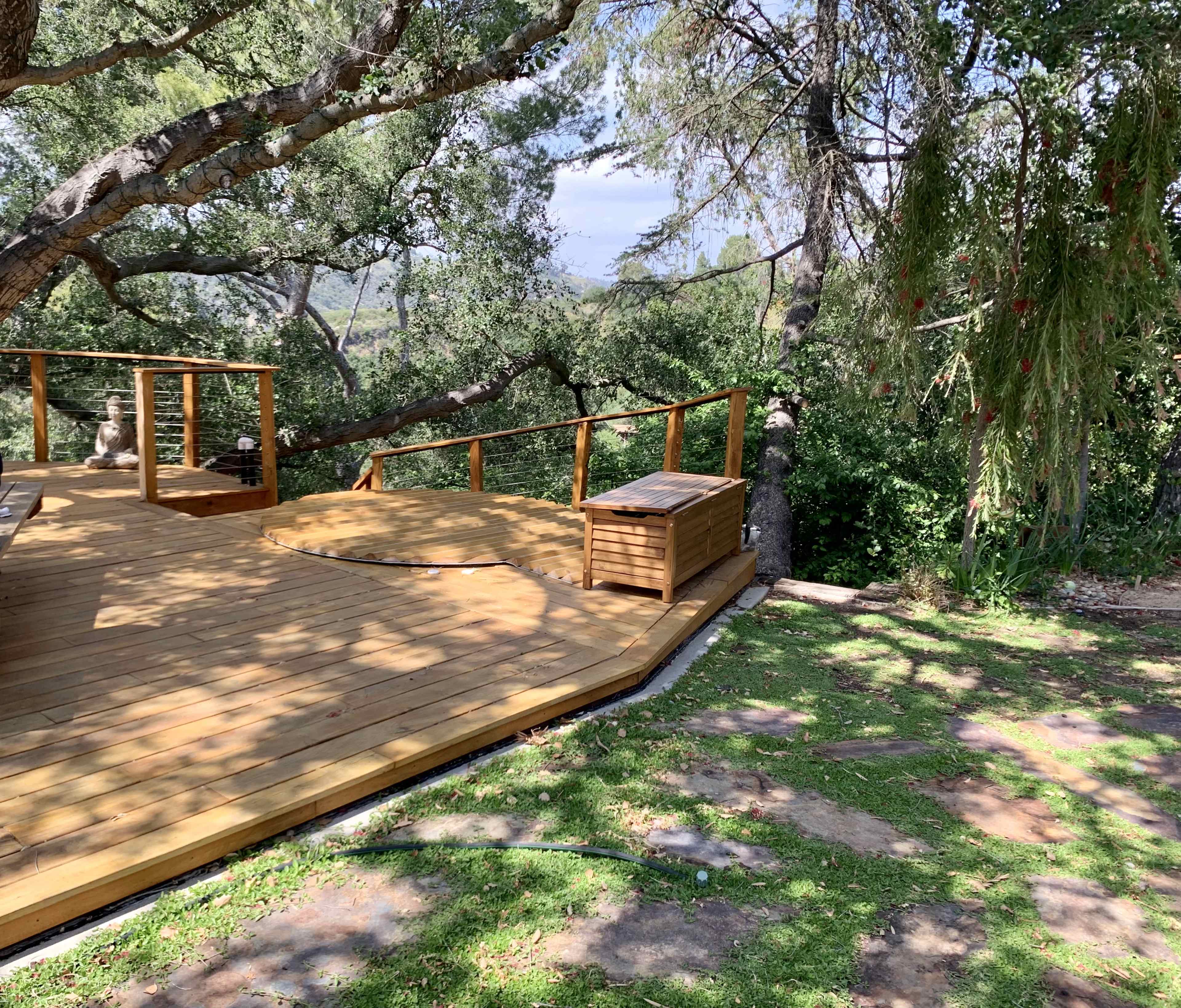 A wooden deck surrounded by trees features a storage box and a grassy area nearby.