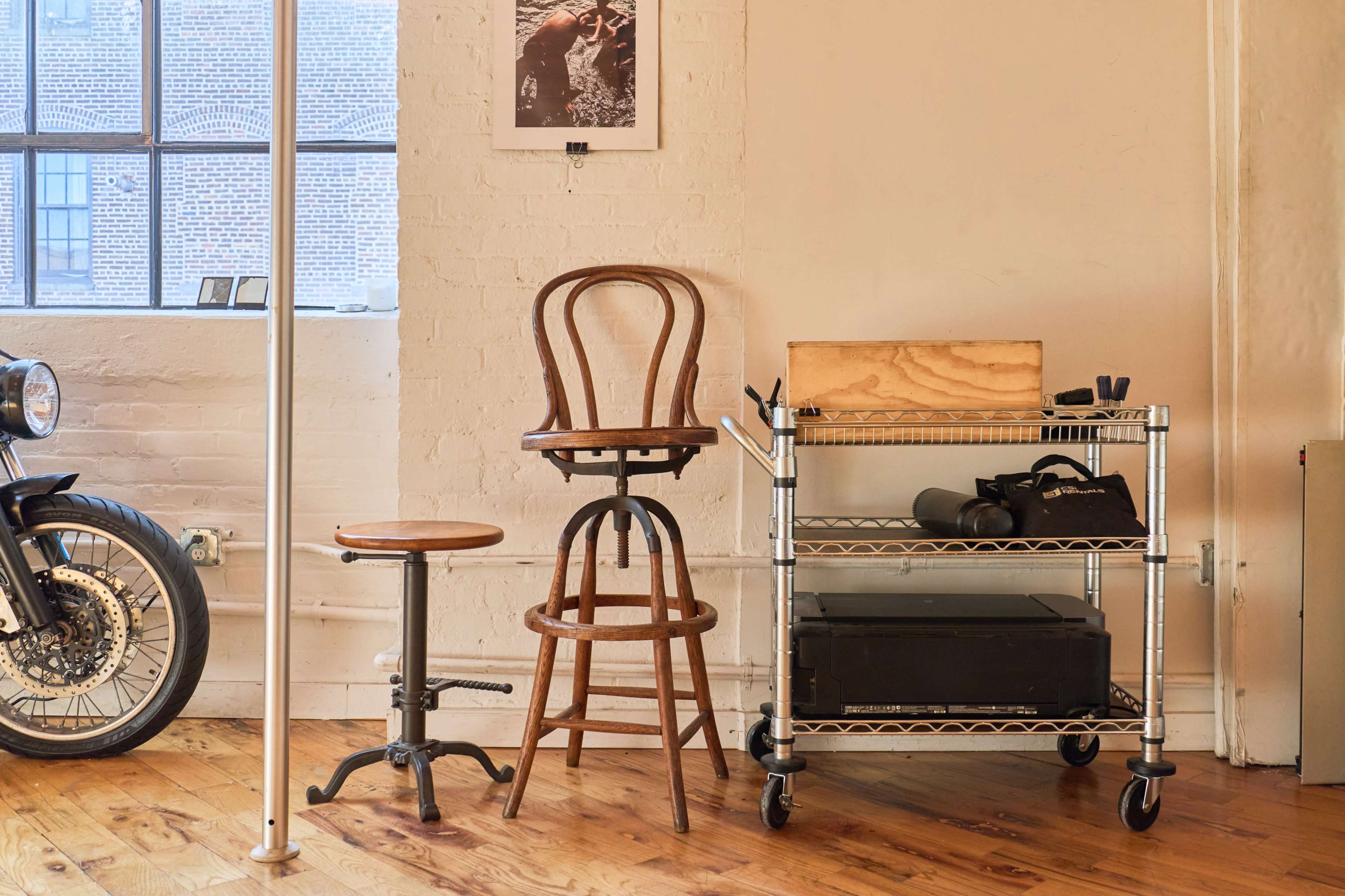 The image shows a wooden high stool and a shorter stool next to a metal rolling cart filled with various items in a room with wood flooring and a large window.