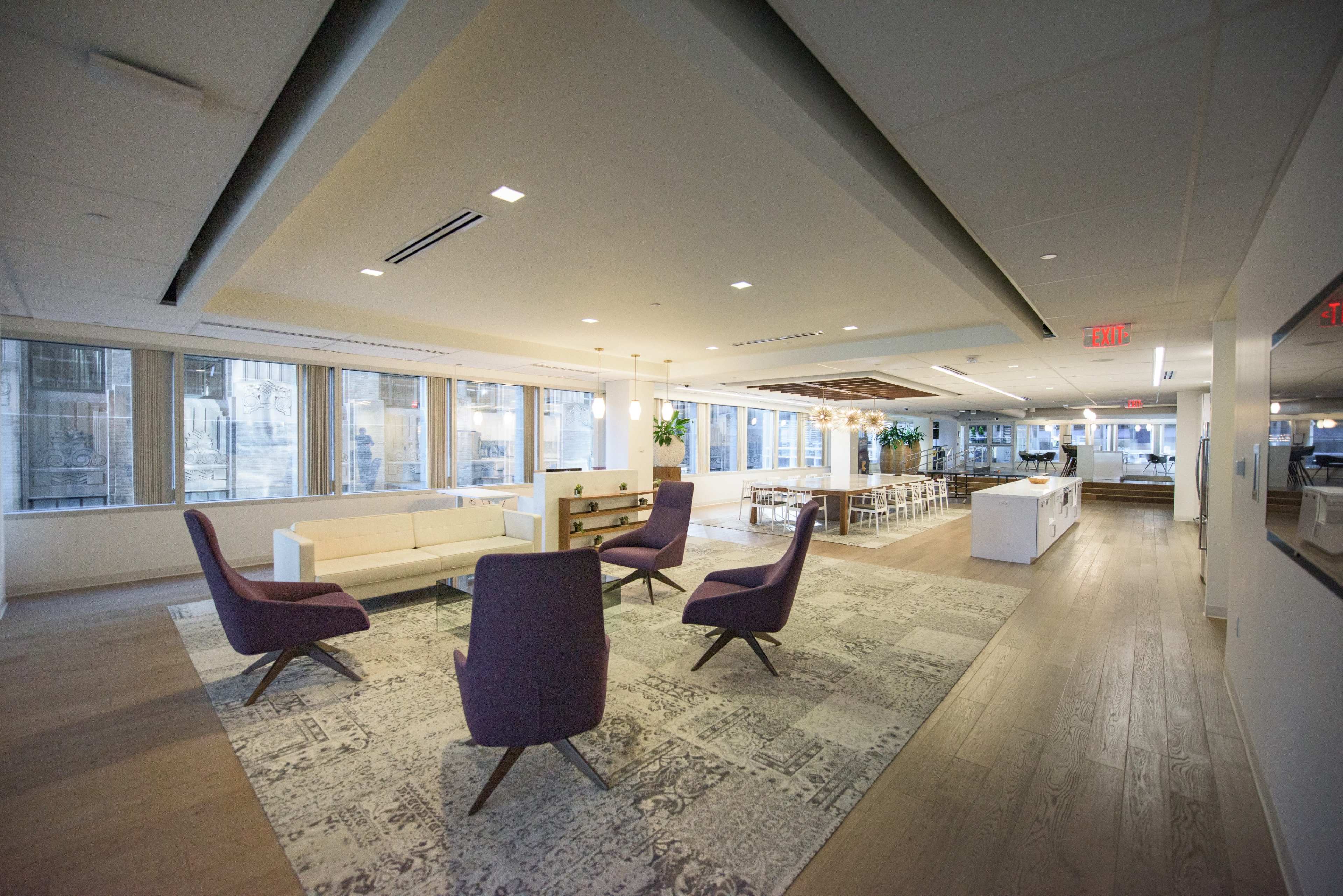 The image features a modern office space with seating areas including purple chairs, a white sofa, and dining tables, illuminated by natural light from large windows.