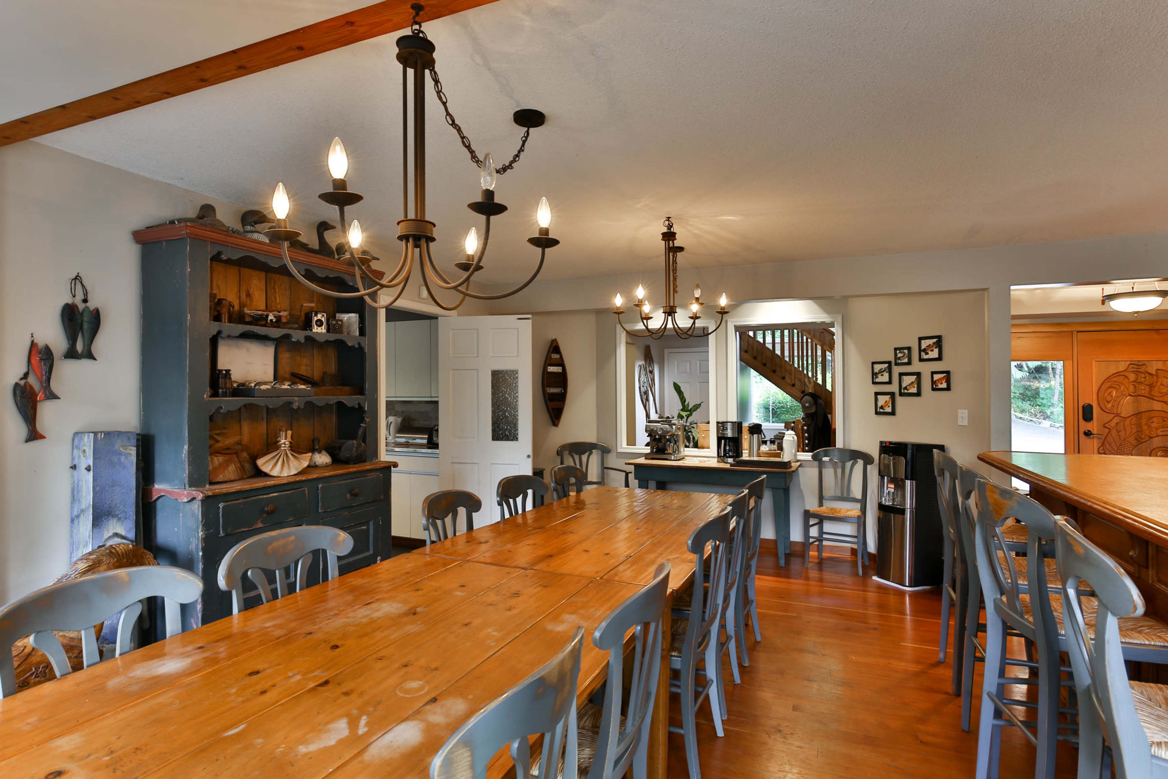 A spacious dining area with a long wooden table surrounded by chairs, accented by two stylish chandeliers and a sideboard filled with decorative items.