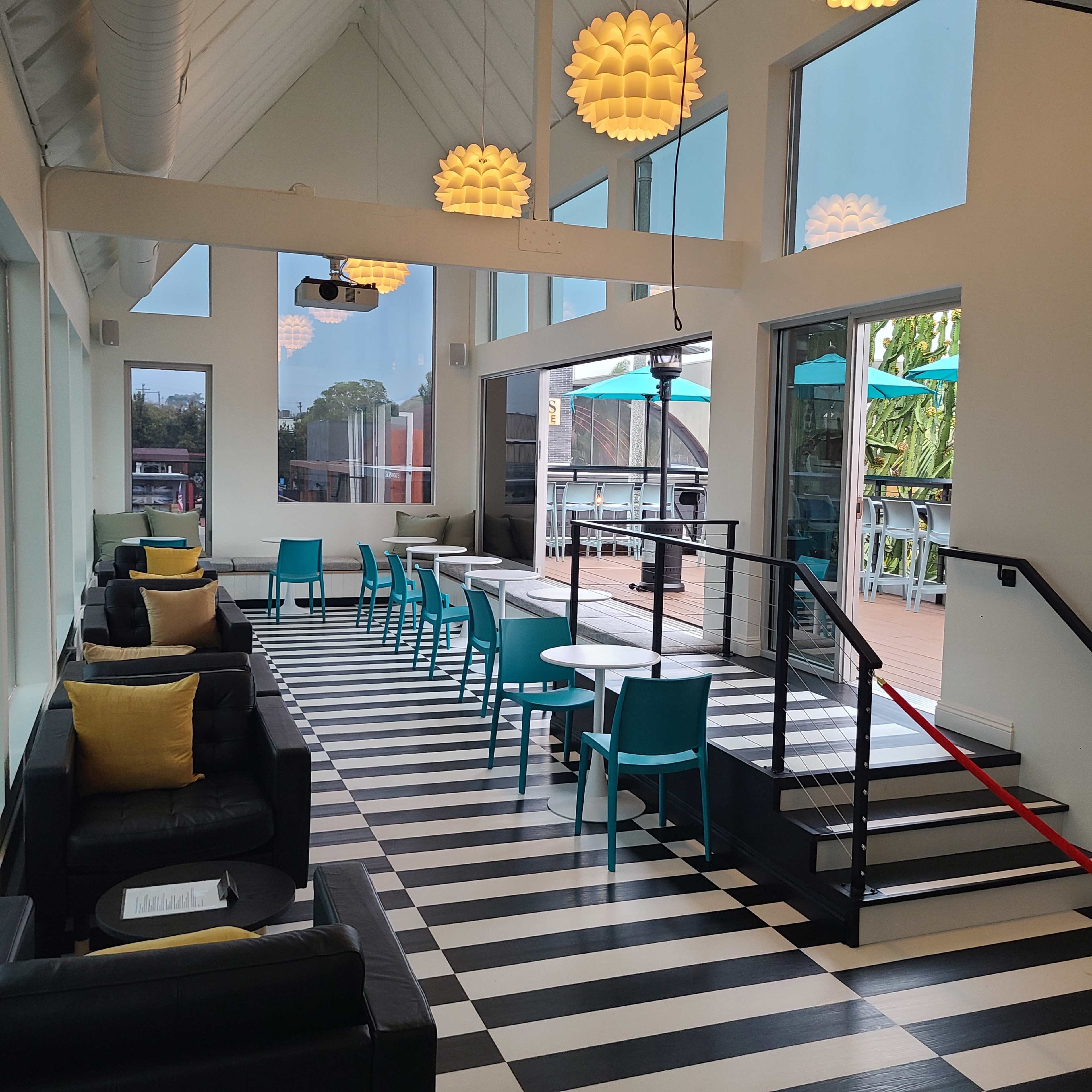 The image shows a modern indoor space with black and white striped flooring, seating areas with black sofas and cushions, and turquoise chairs arranged along a dining area with large windows.