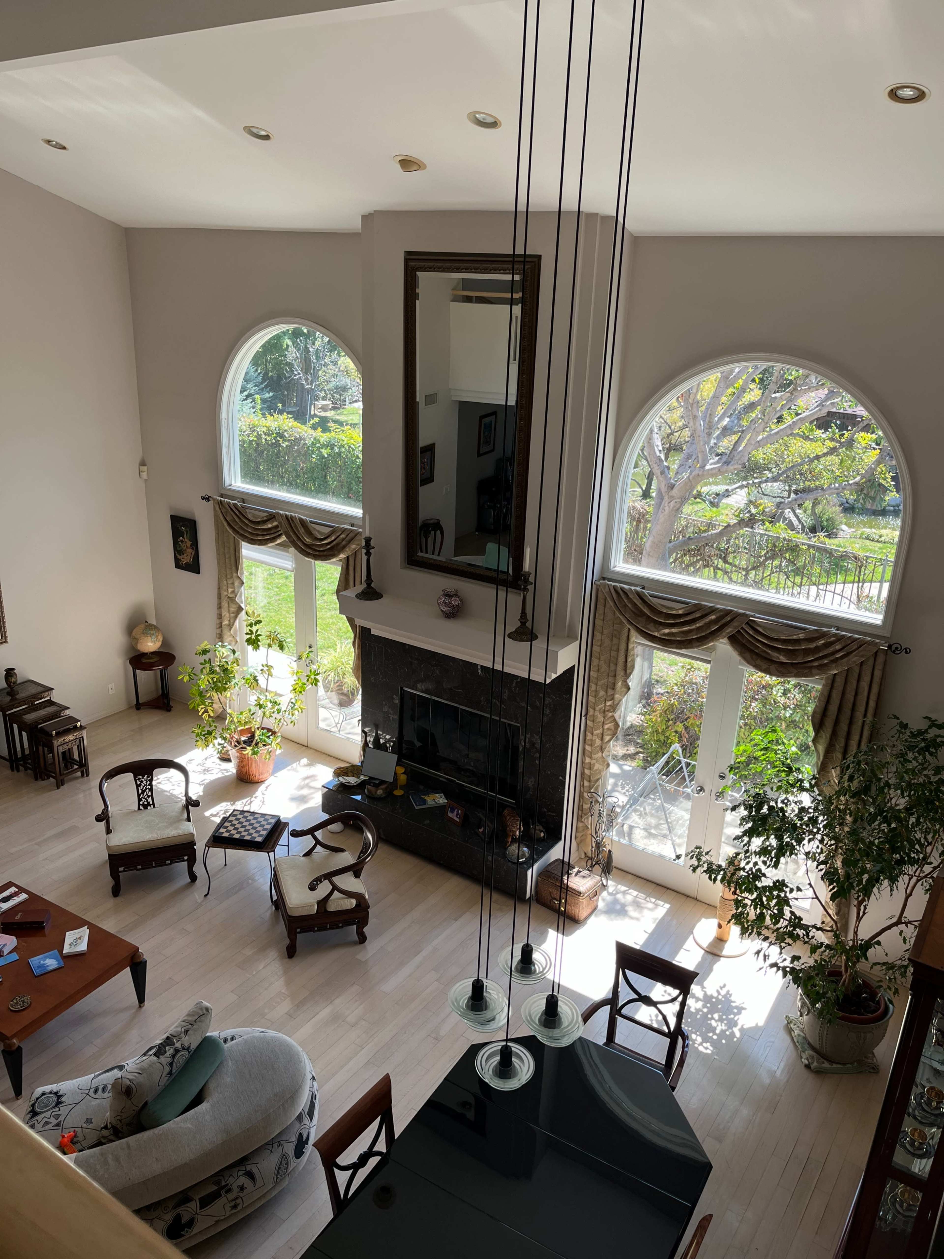 The image shows a spacious living room with large arched windows, a fireplace, and various plants, as viewed from an upper level.