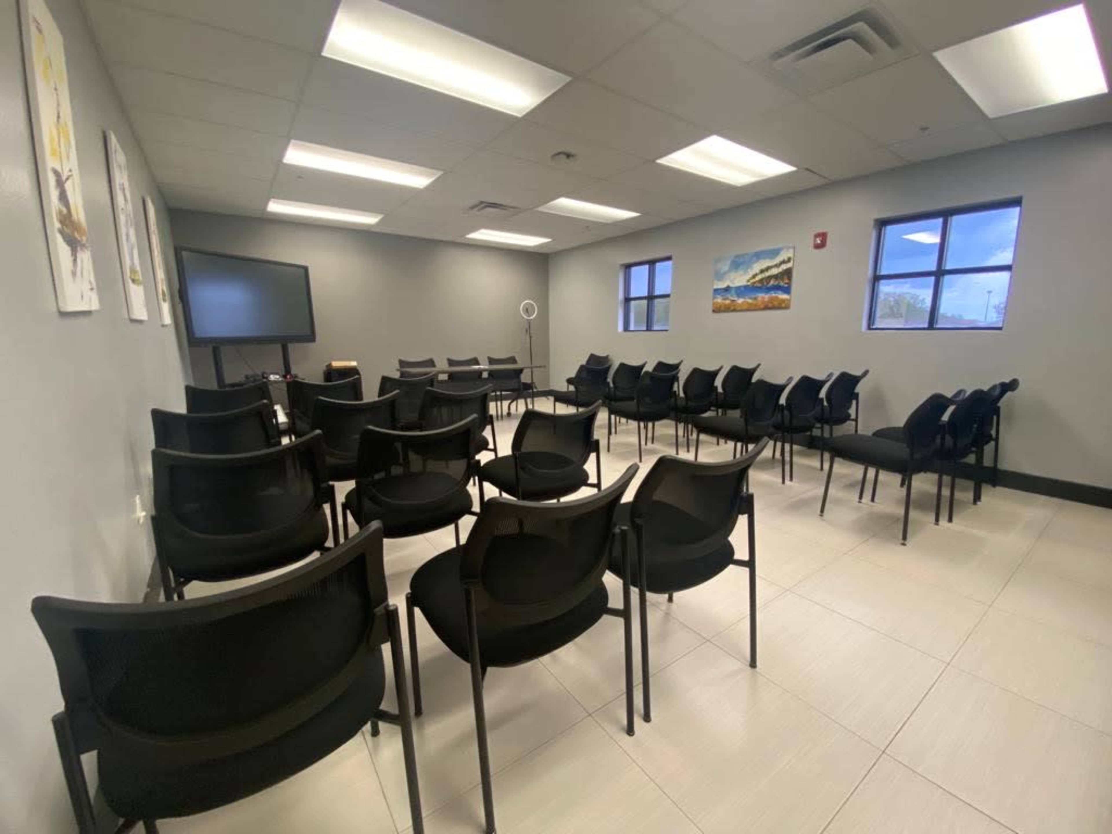 The image shows a meeting room with rows of black chairs arranged around a central space, a television on a stand, and artwork on the walls.