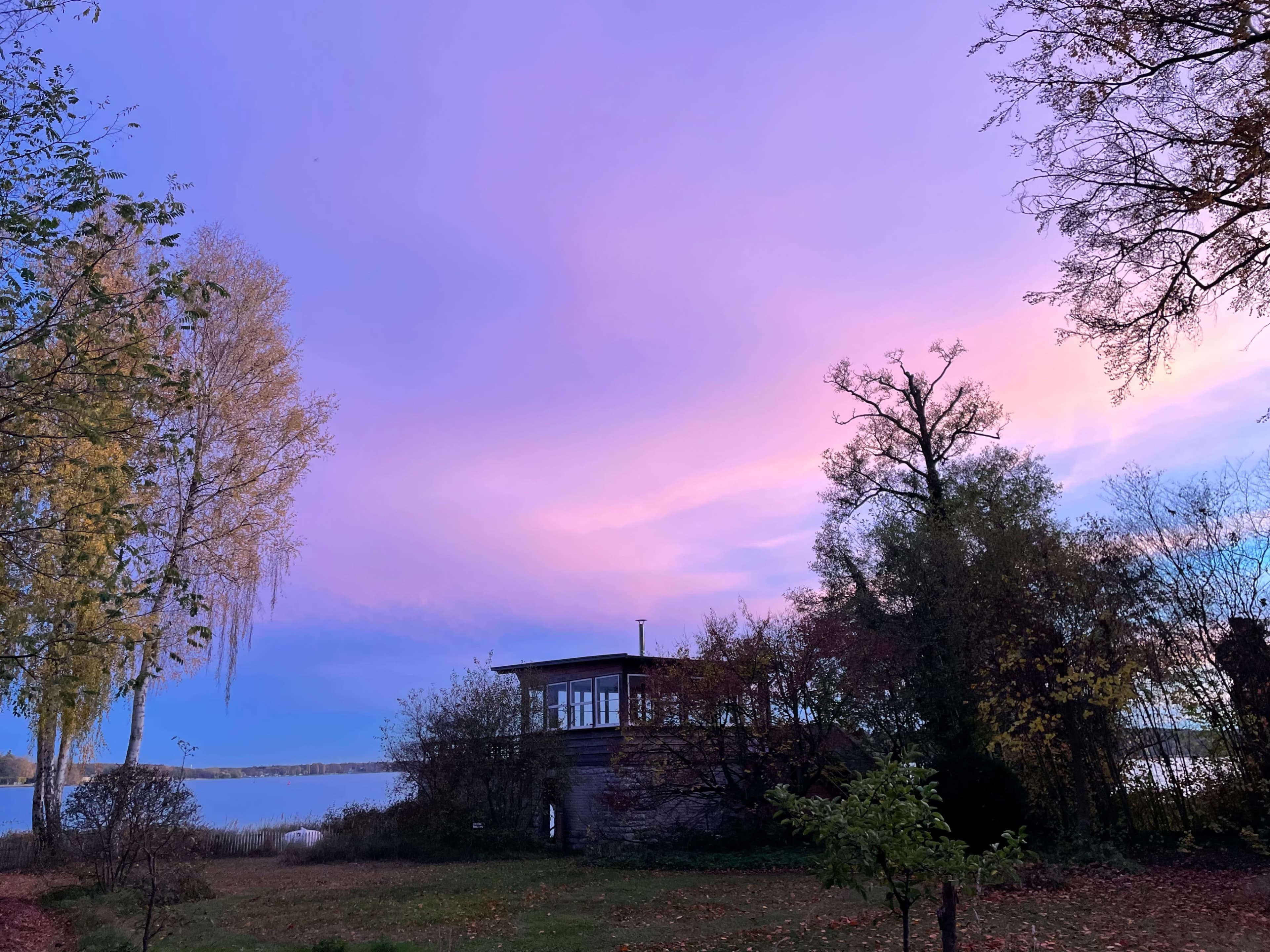 A wooden building sits beside a calm lake under a colorful sky during twilight.