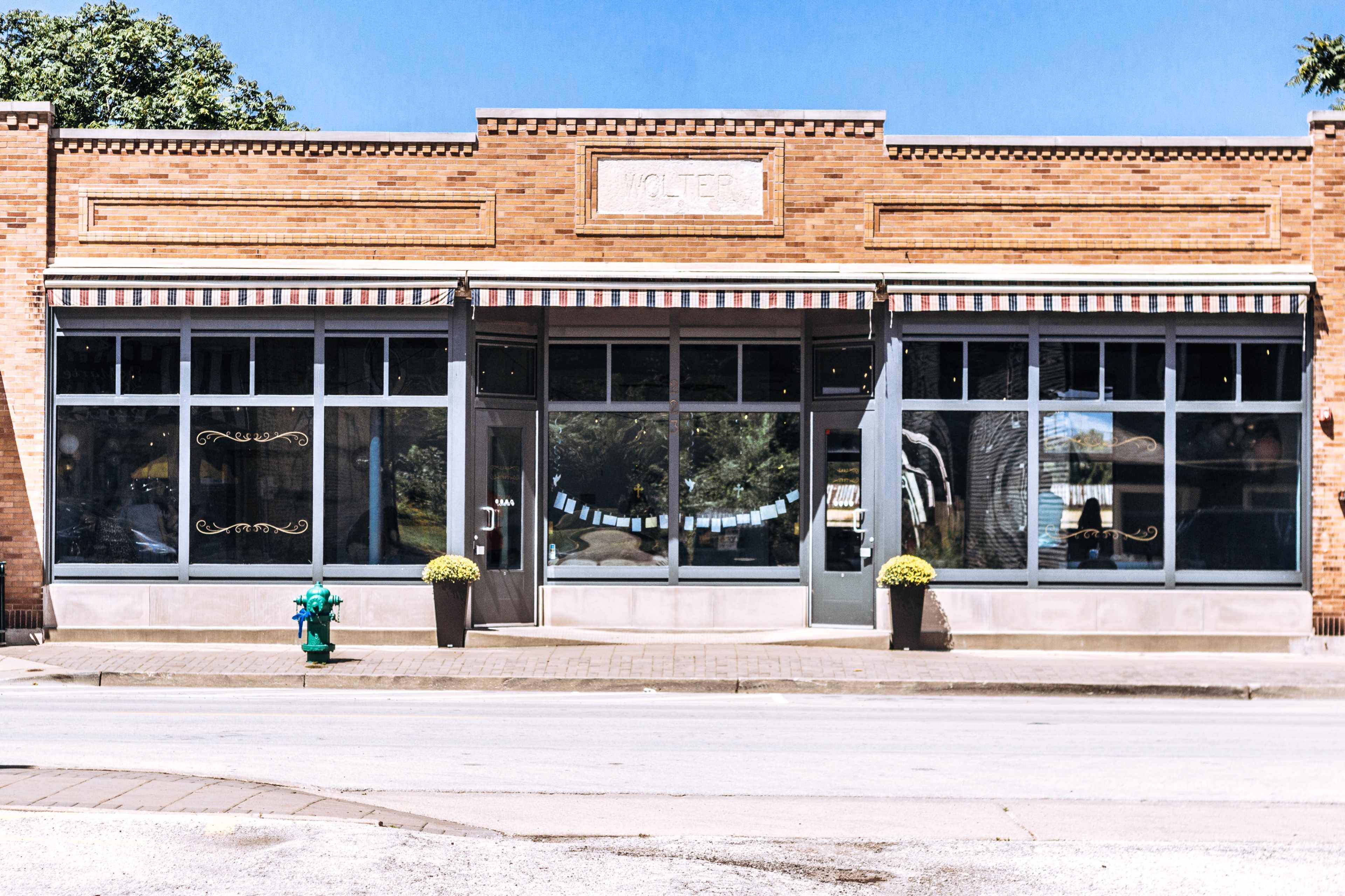 The image shows a brick storefront with large glass windows and striped awnings, featuring planters with yellow flowers on either side of the entrance.