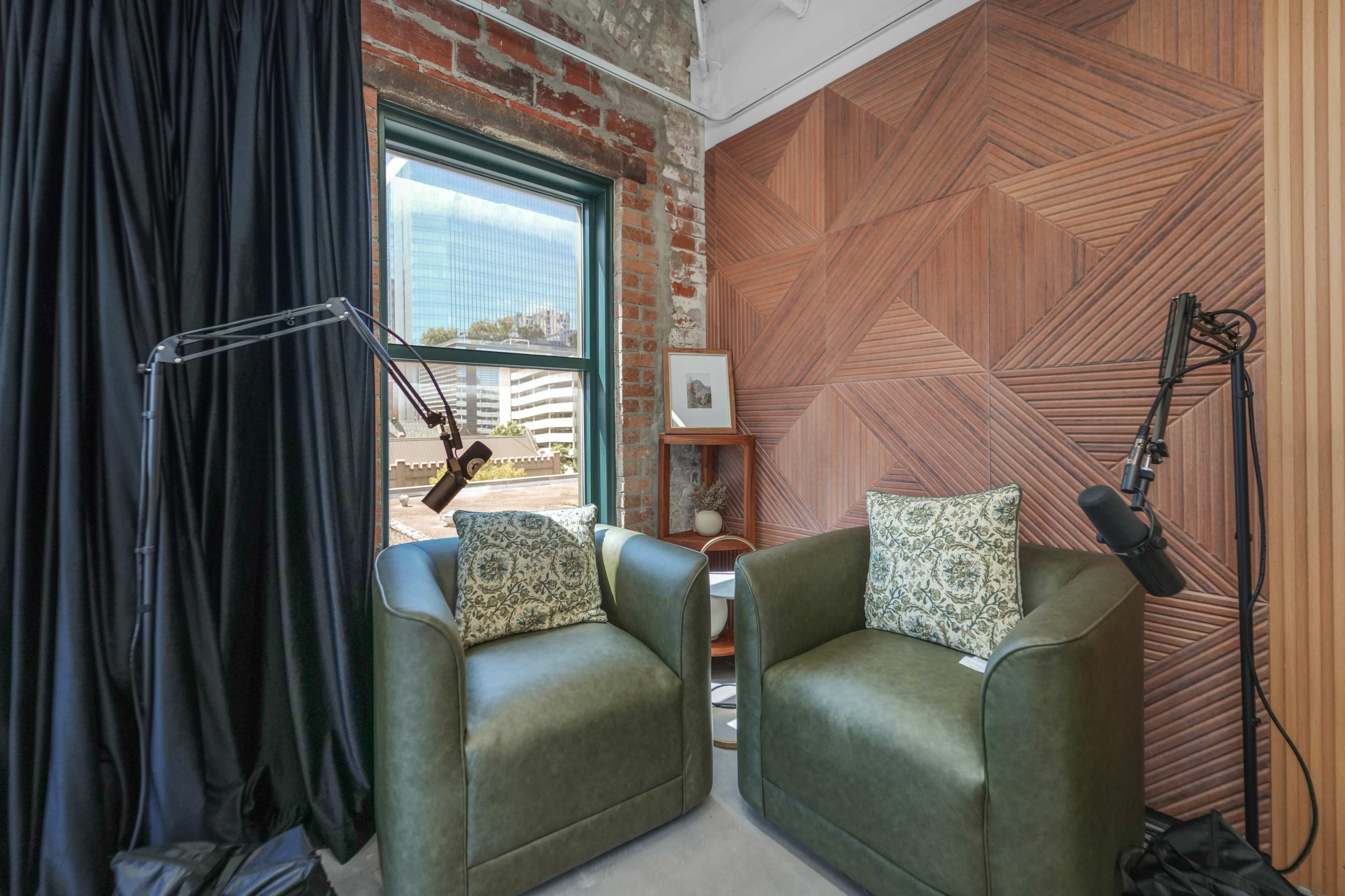 The image shows a corner of a room featuring two green armchairs with patterned cushions, next to a window and a geometric wood accent wall.