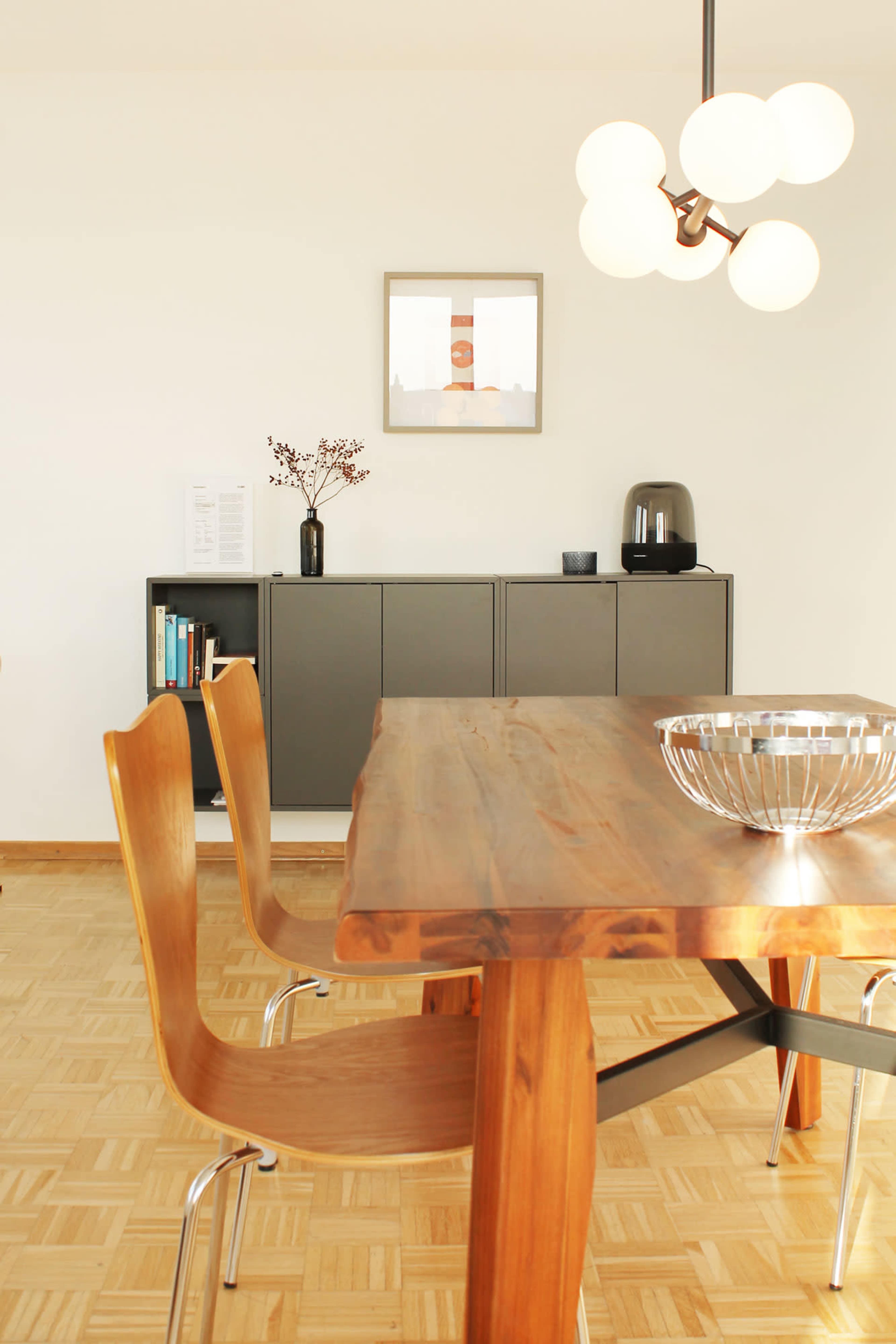 A wooden dining table with two chairs sits in a minimally furnished room featuring a light fixture, a framed picture on the wall, and a modern sideboard.