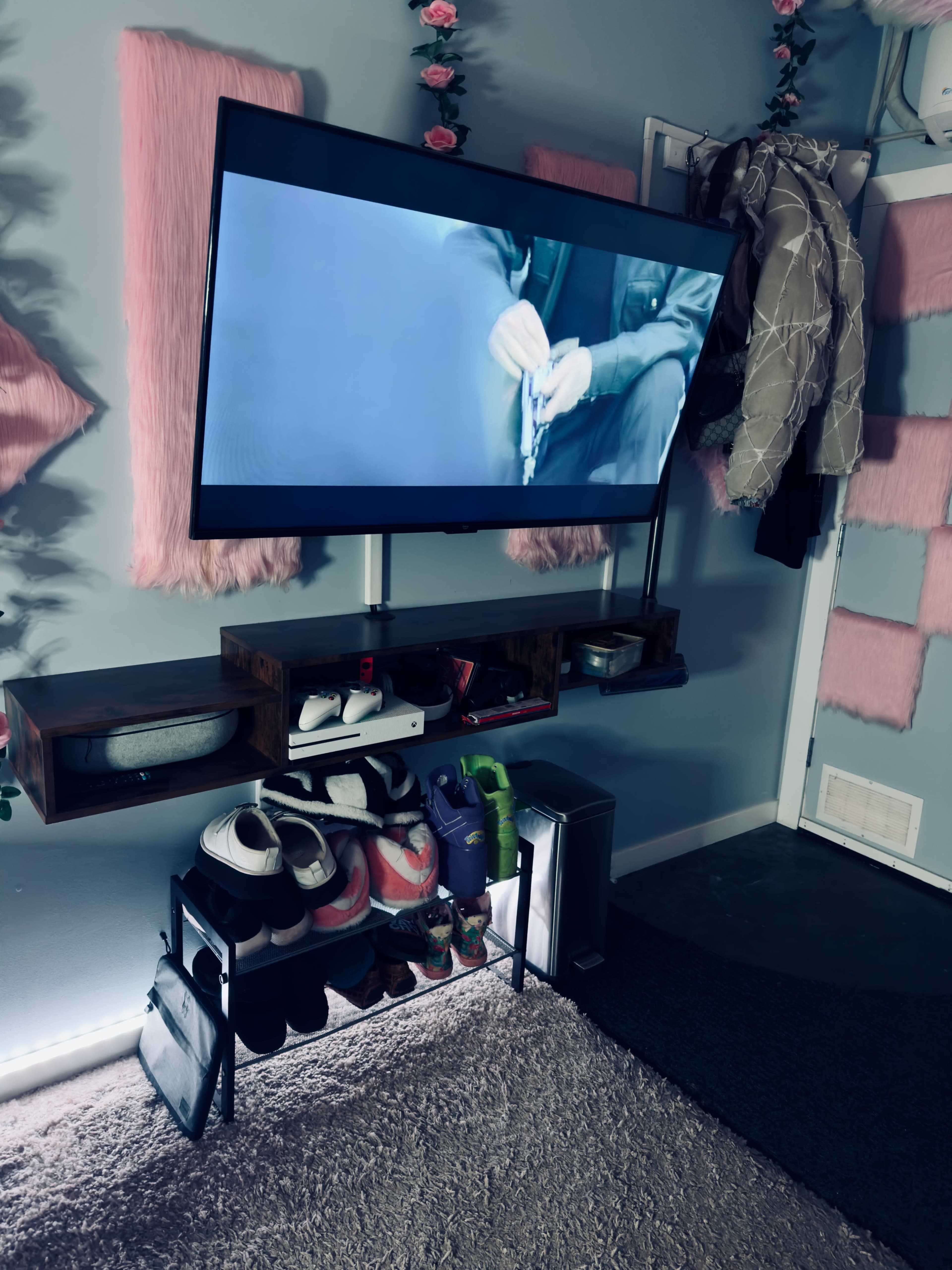 A living space with a mounted television displaying a scene, a shelf holding shoes, and a wall decorated with pink panels and a coat hanging nearby.