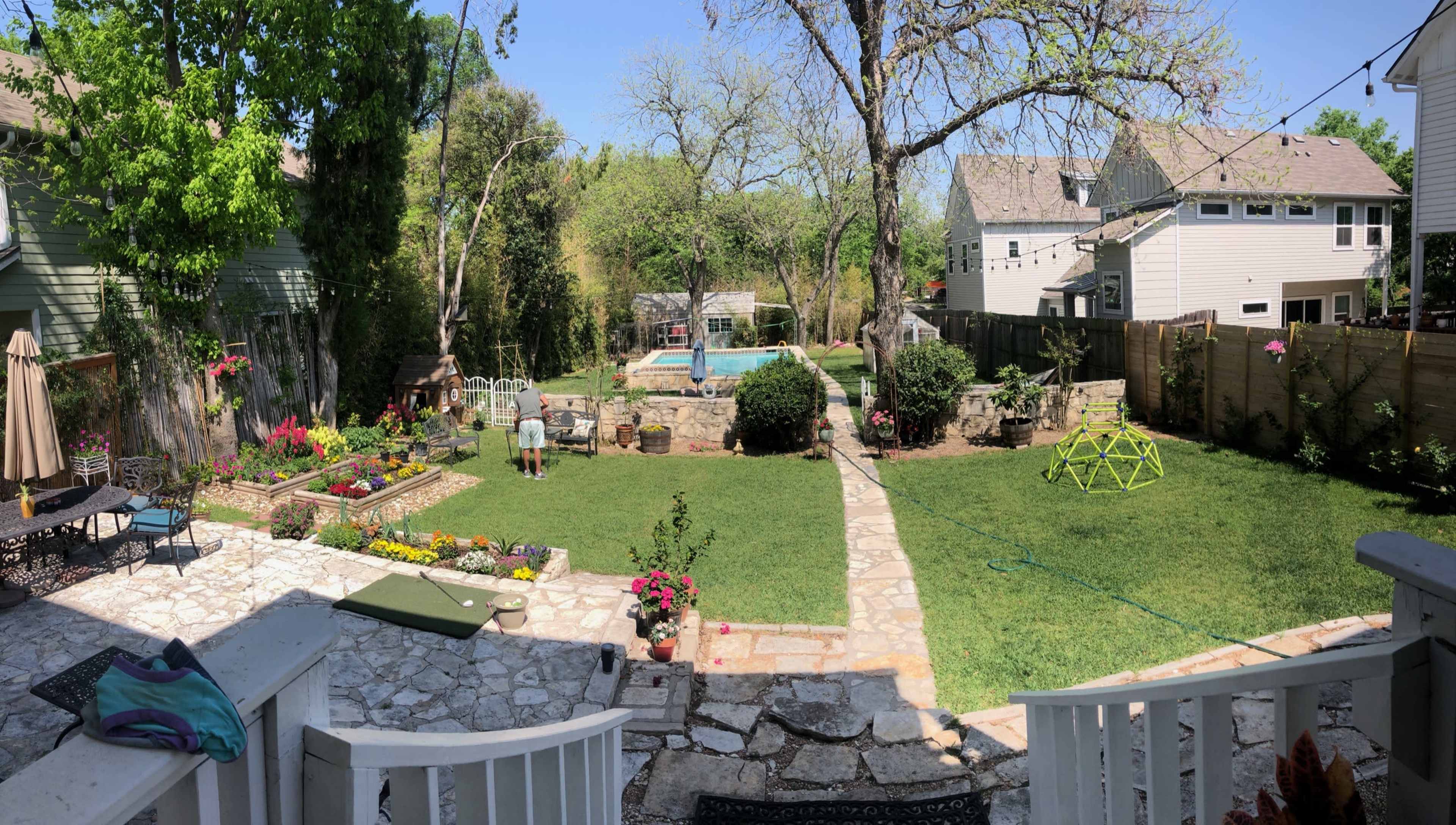 A backyard scene features a grassy area with a swimming pool, stone landscaping, flower beds, and a person tending to the garden.