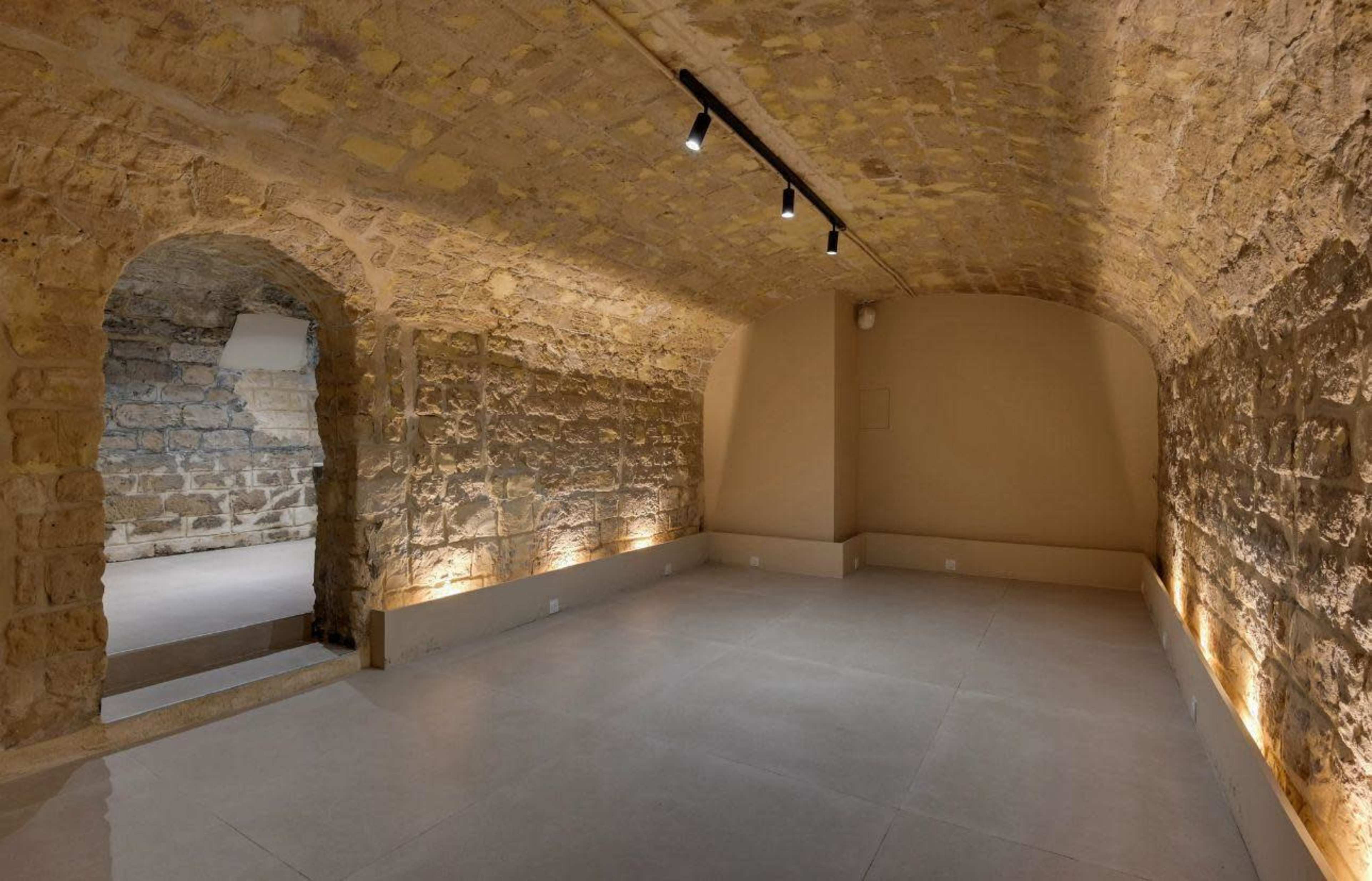 The image shows a spacious, well-lit room with stone walls and a vaulted ceiling, featuring a minimalist design and concrete flooring.