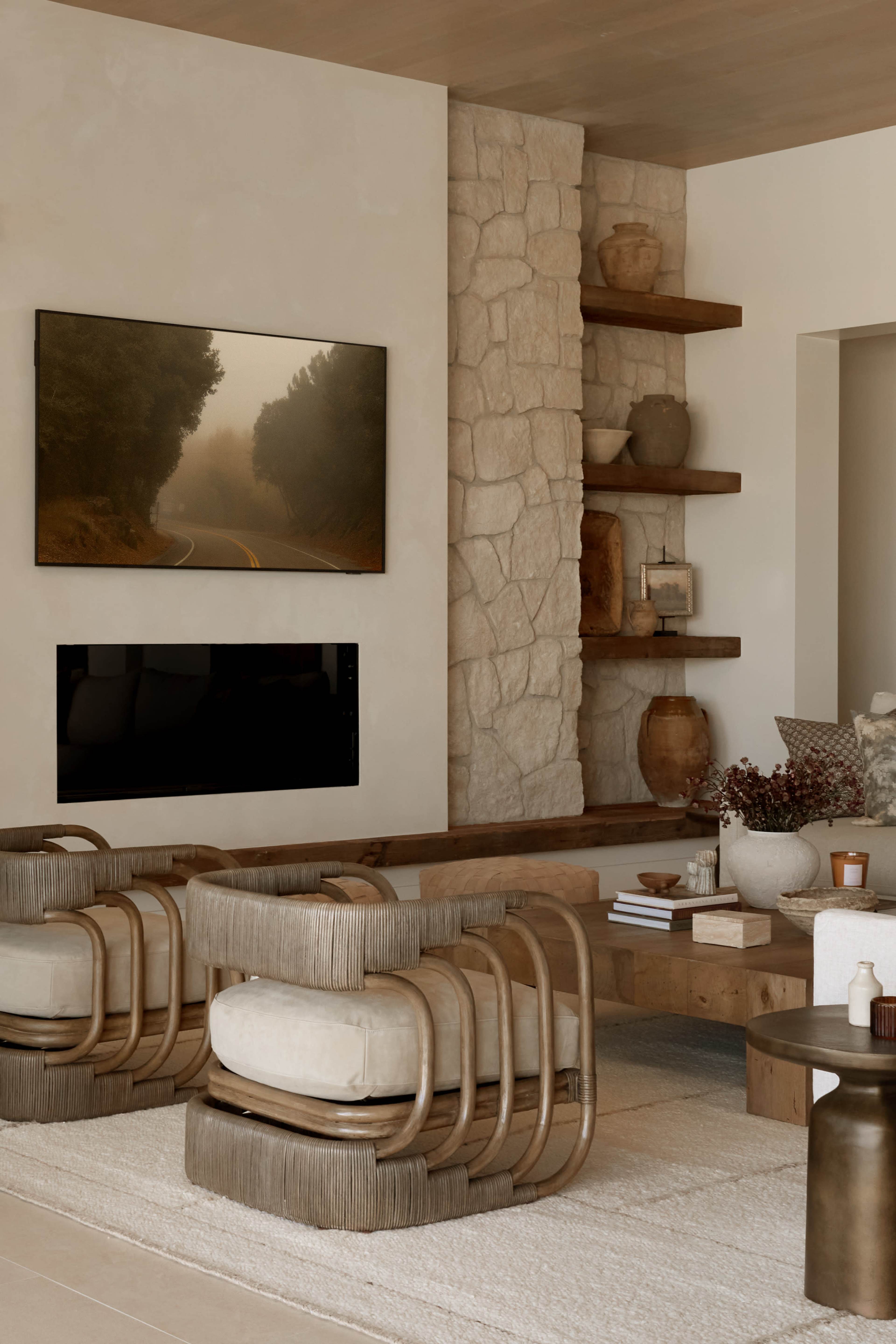 The image shows a modern living room with two curved armchairs, a stone wall, shelves displaying decorative items, and a television mounted above a low wooden table.