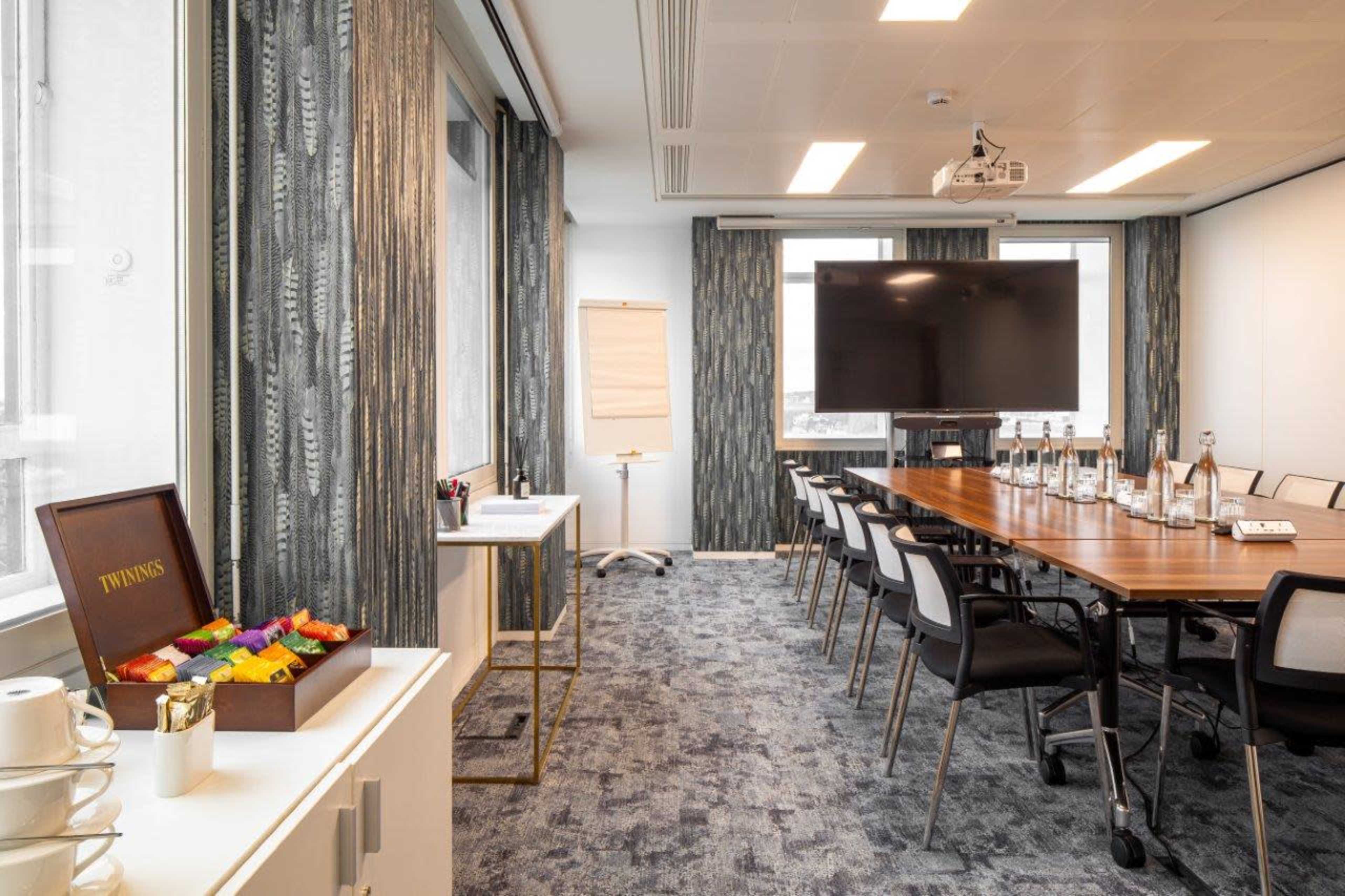 A modern conference room features a long wooden table set with glass water bottles, a large screen at one end, and a flip chart on a stand.