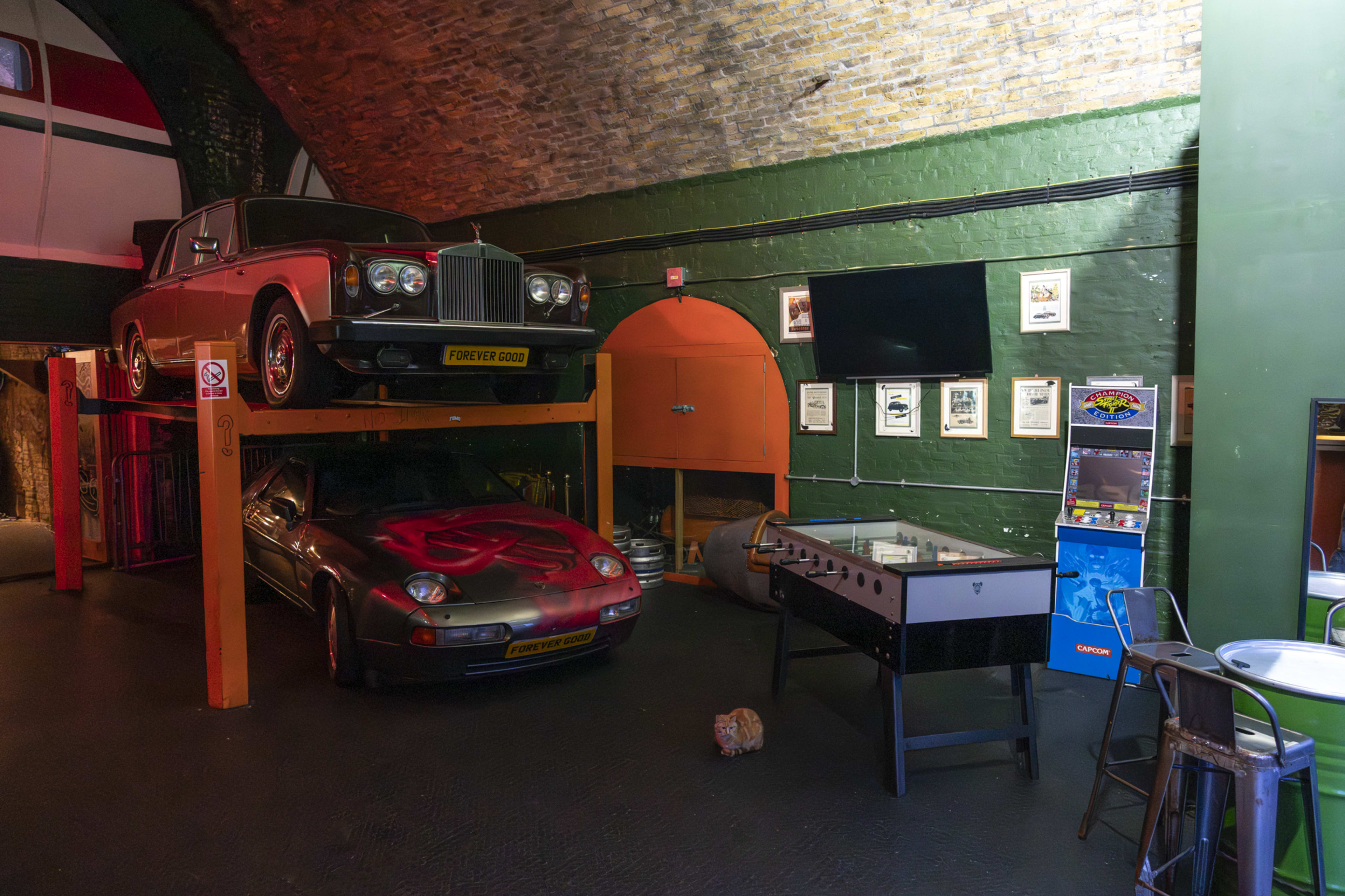 The image shows a unique interior space featuring a classic car displayed on a raised platform above another car, with a foosball table and a vending machine visible in the foreground.