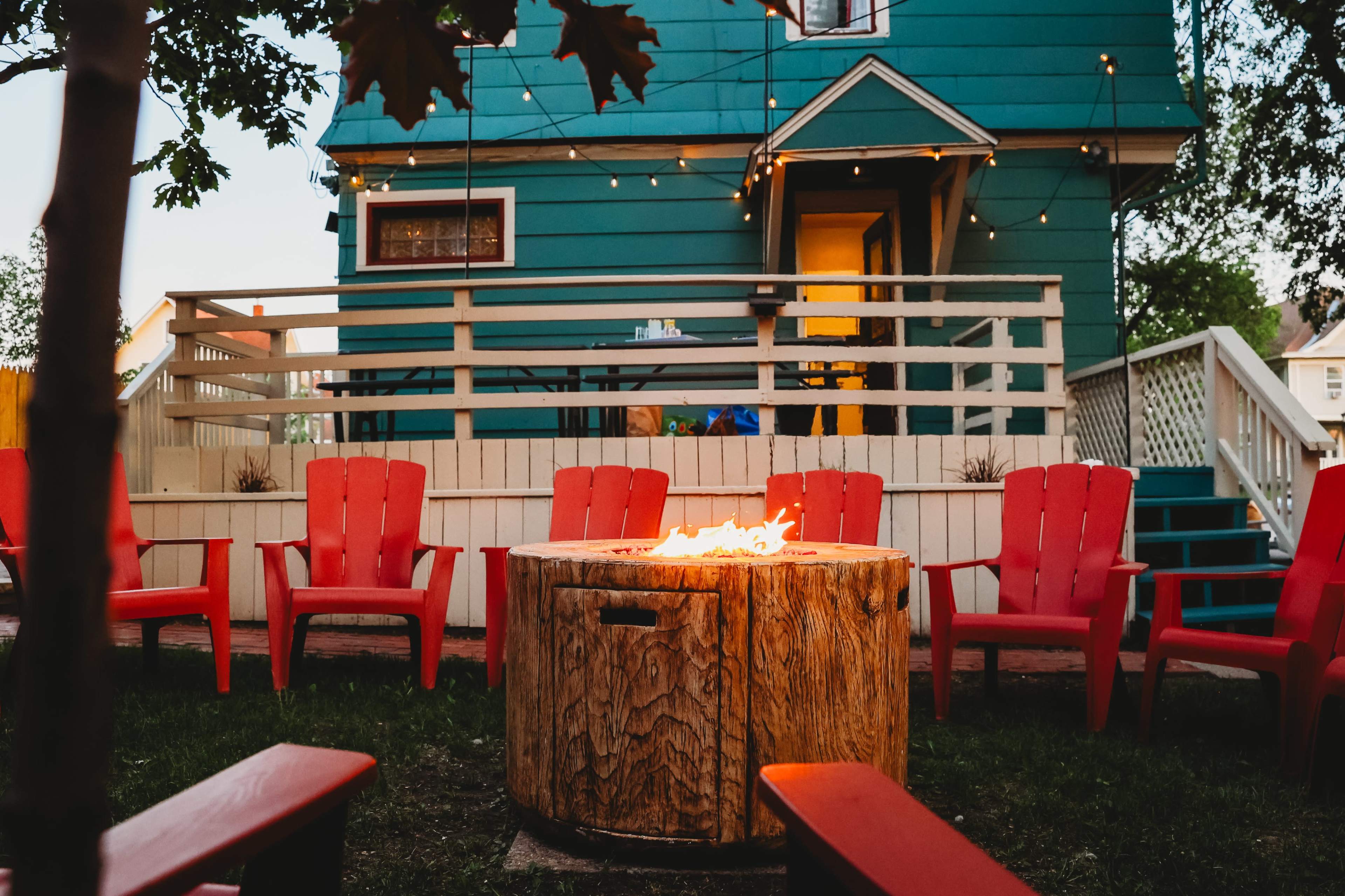 The image shows a circular fire pit surrounded by red Adirondack chairs in a backyard with a teal house in the background and string lights overhead.
