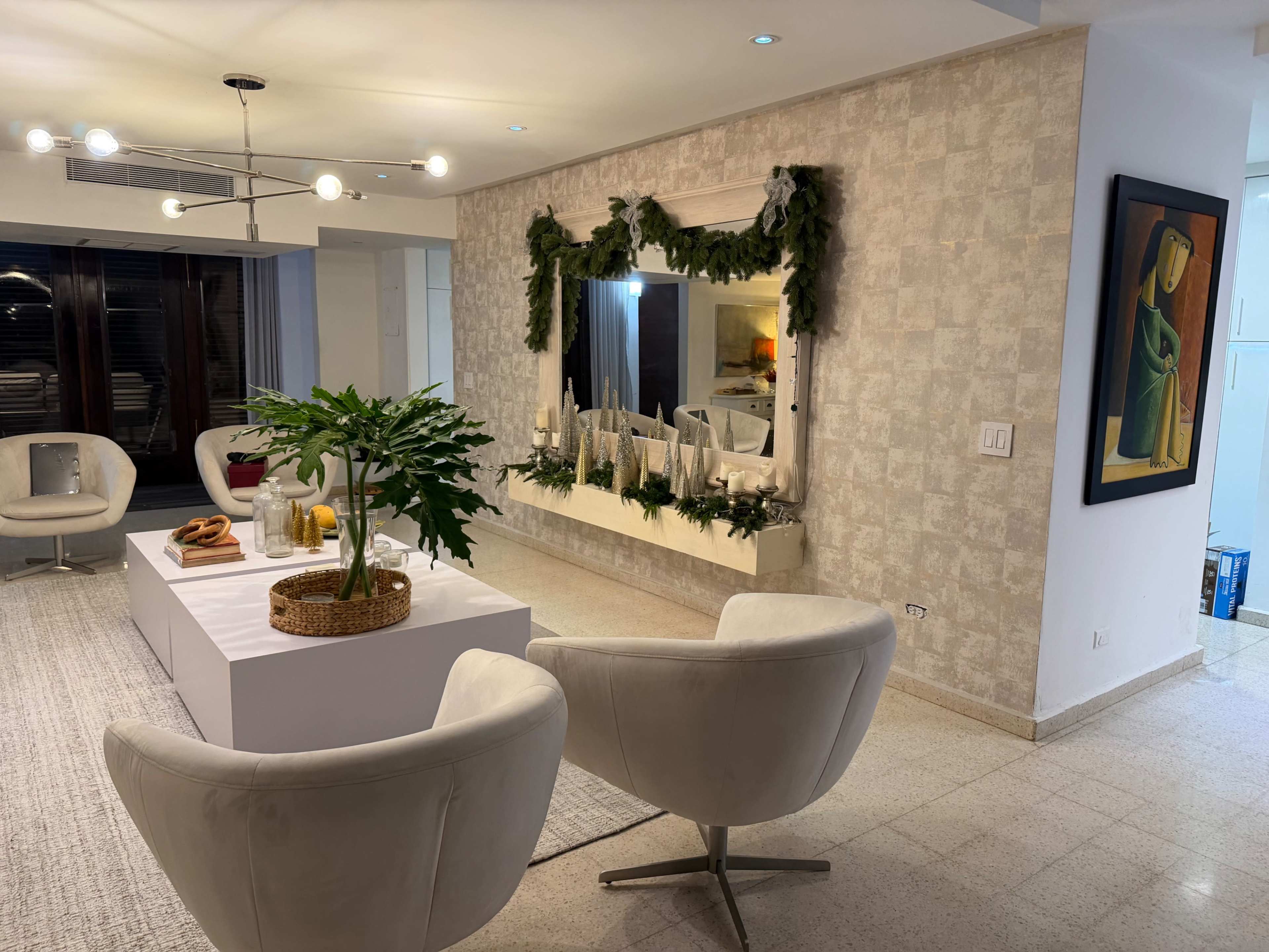 The image shows a modern living room with two white chairs, a central coffee table, and a large mirror decorated with greenery, against a textured light-colored wall.