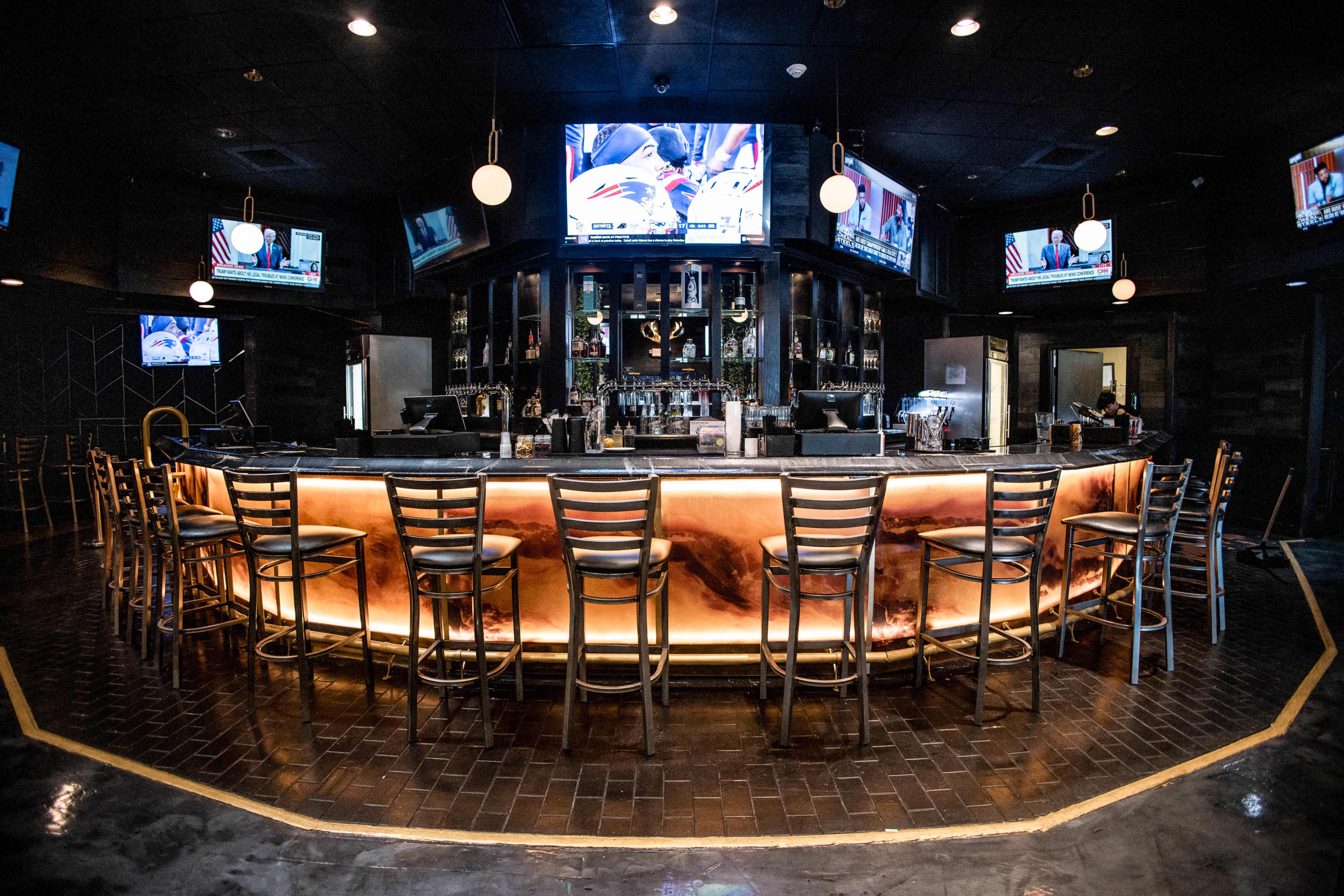 The image shows a dimly lit bar with a circular counter, several tall metal stools, and multiple televisions displaying sports events.