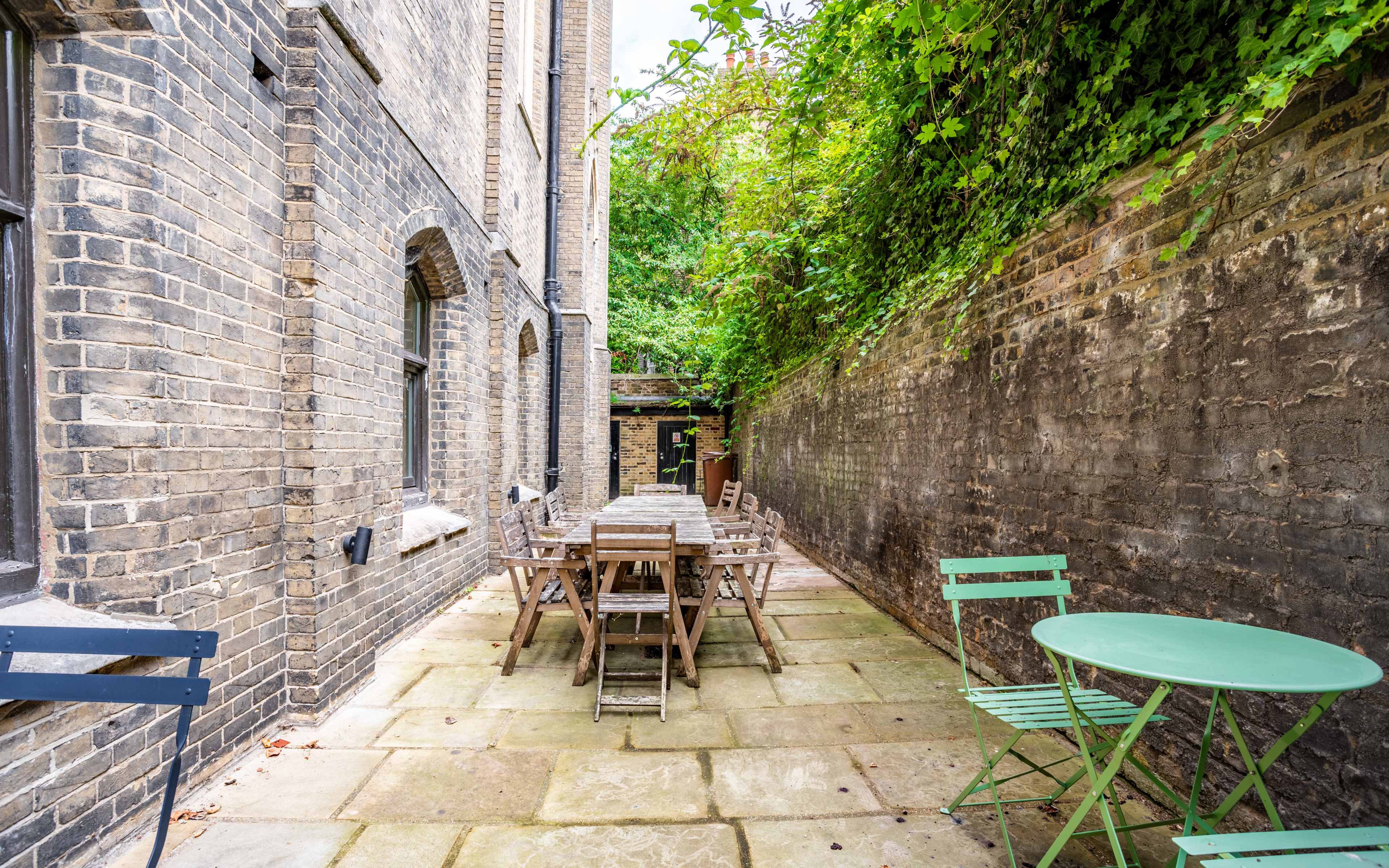 A narrow, paved courtyard features a long wooden dining table surrounded by chairs, flanked by brick walls and overgrown greenery.