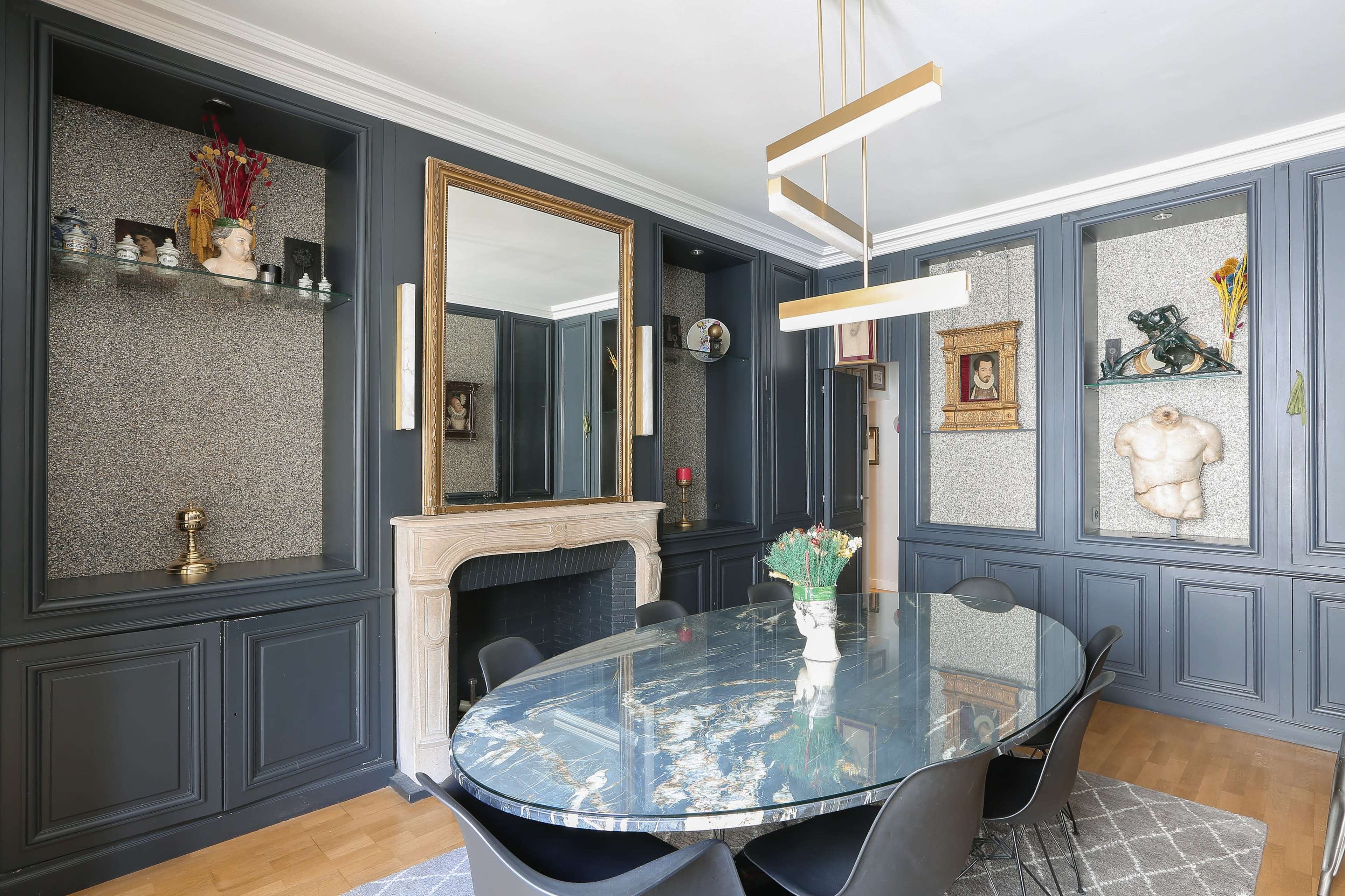 The image shows a modern dining room featuring a round glass table surrounded by black chairs, with framed art and decorative objects displayed on the walls.