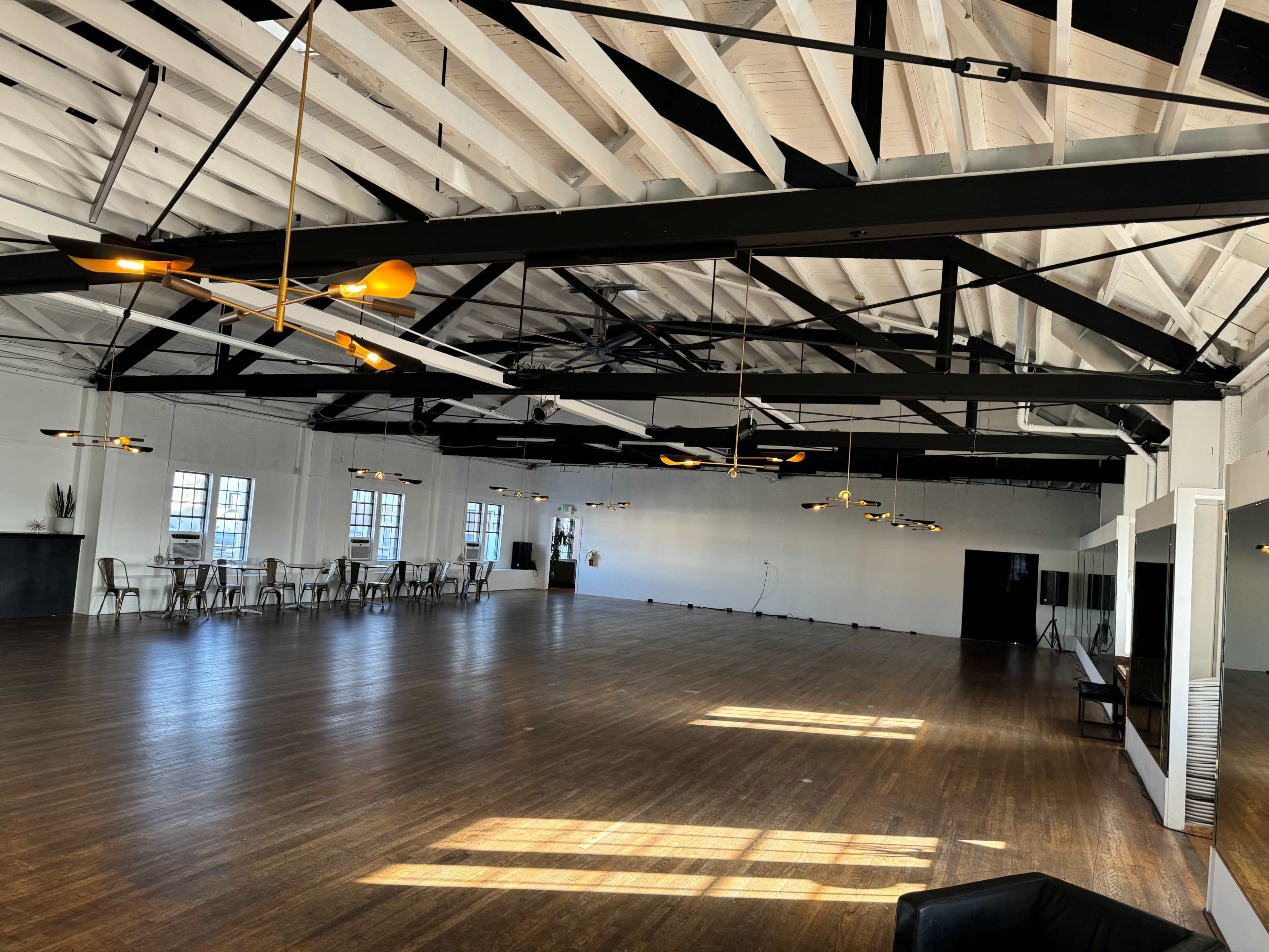 The image shows a spacious, well-lit event space with wooden flooring, large windows, and contemporary lighting fixtures.