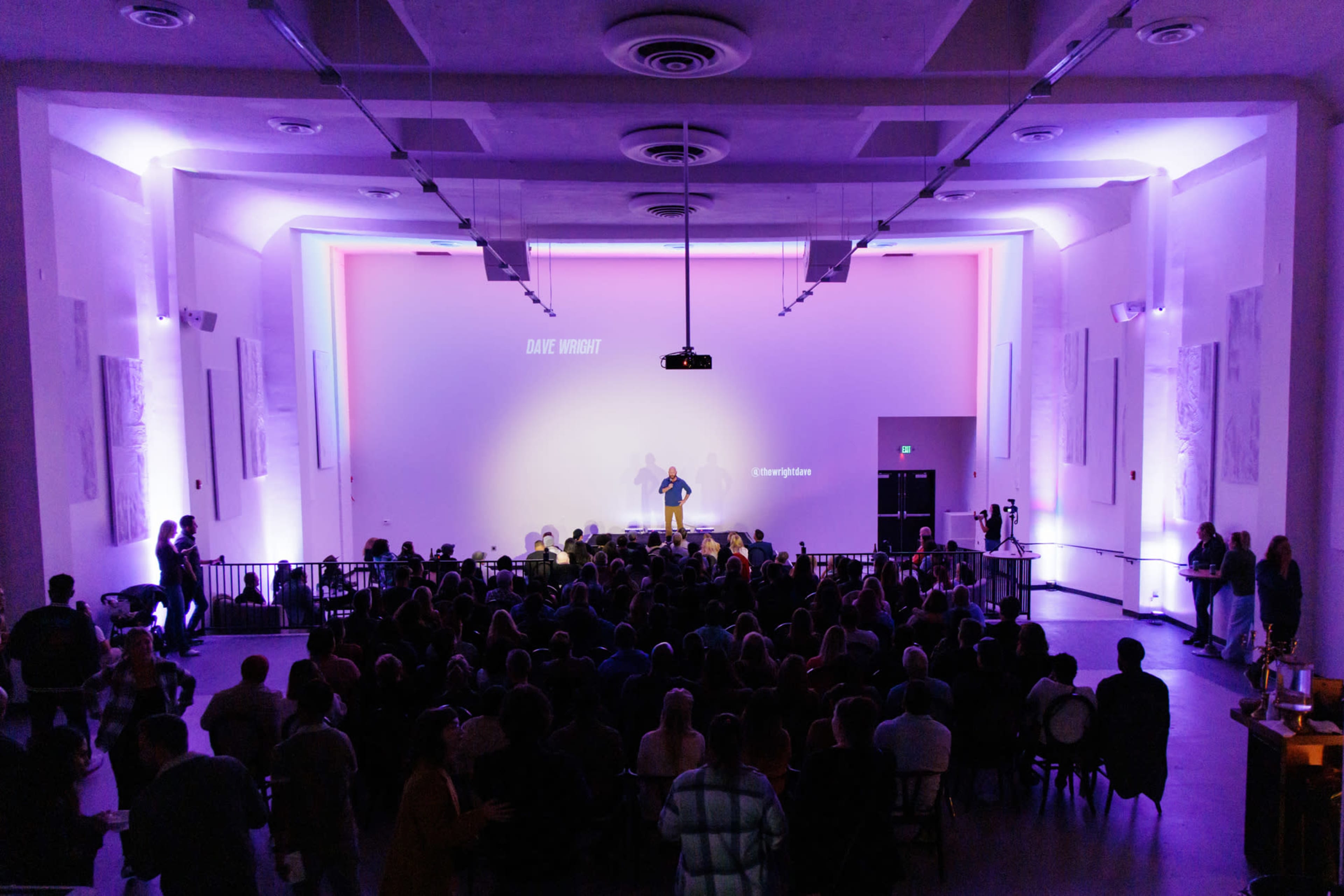 A speaker stands on a stage in a well-lit auditorium filled with an audience.