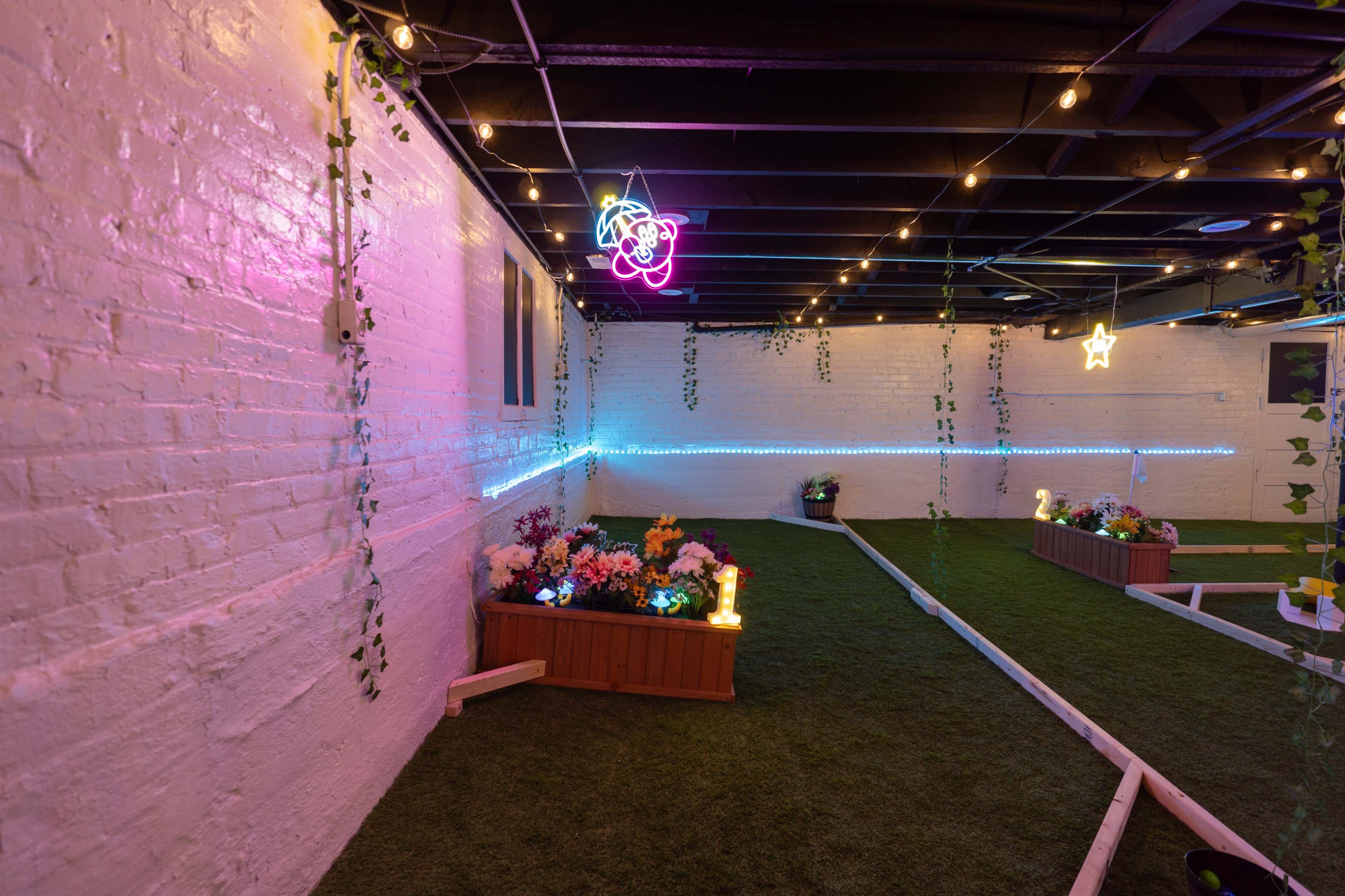 An indoor space with artificial grass, flower boxes, and colorful neon lights hanging from the ceiling.
